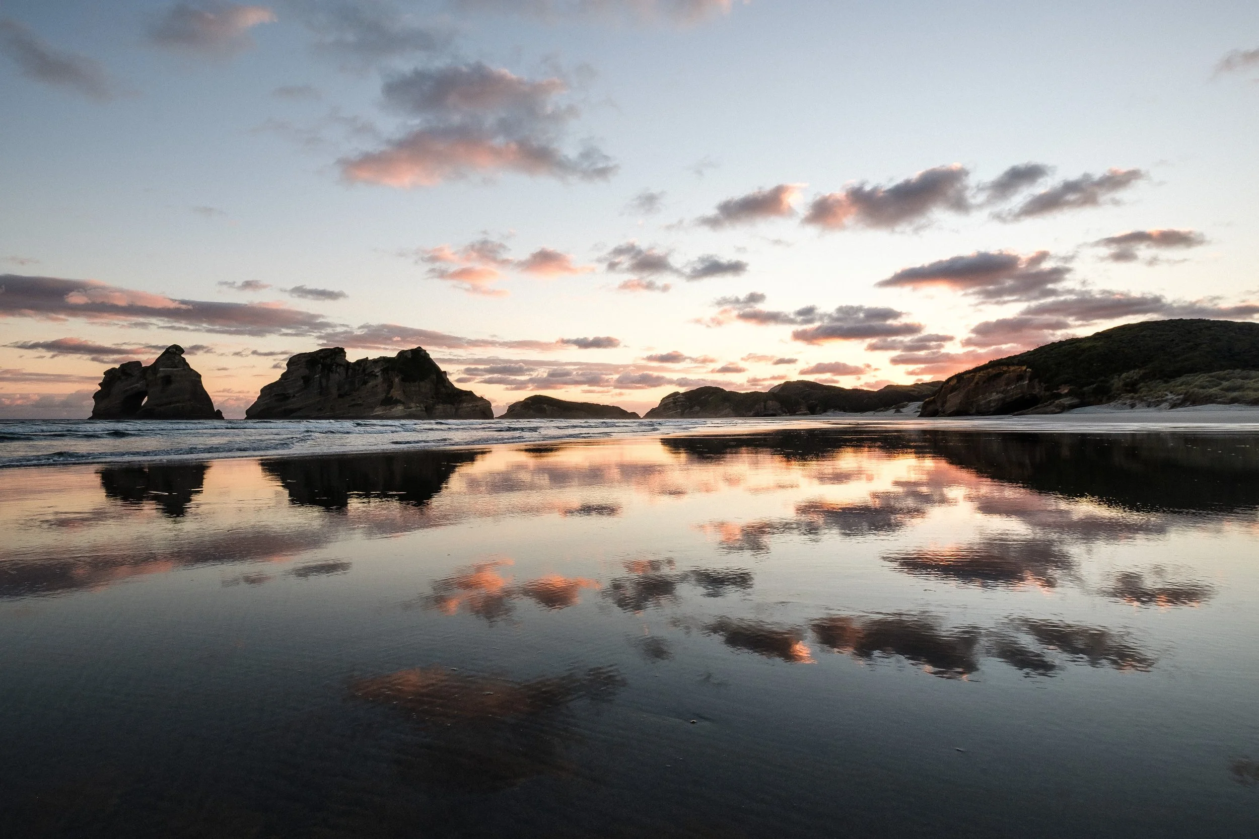 Sunset over a beach with rocks and cliffs, calm water reflecting the colorful sky and clouds.