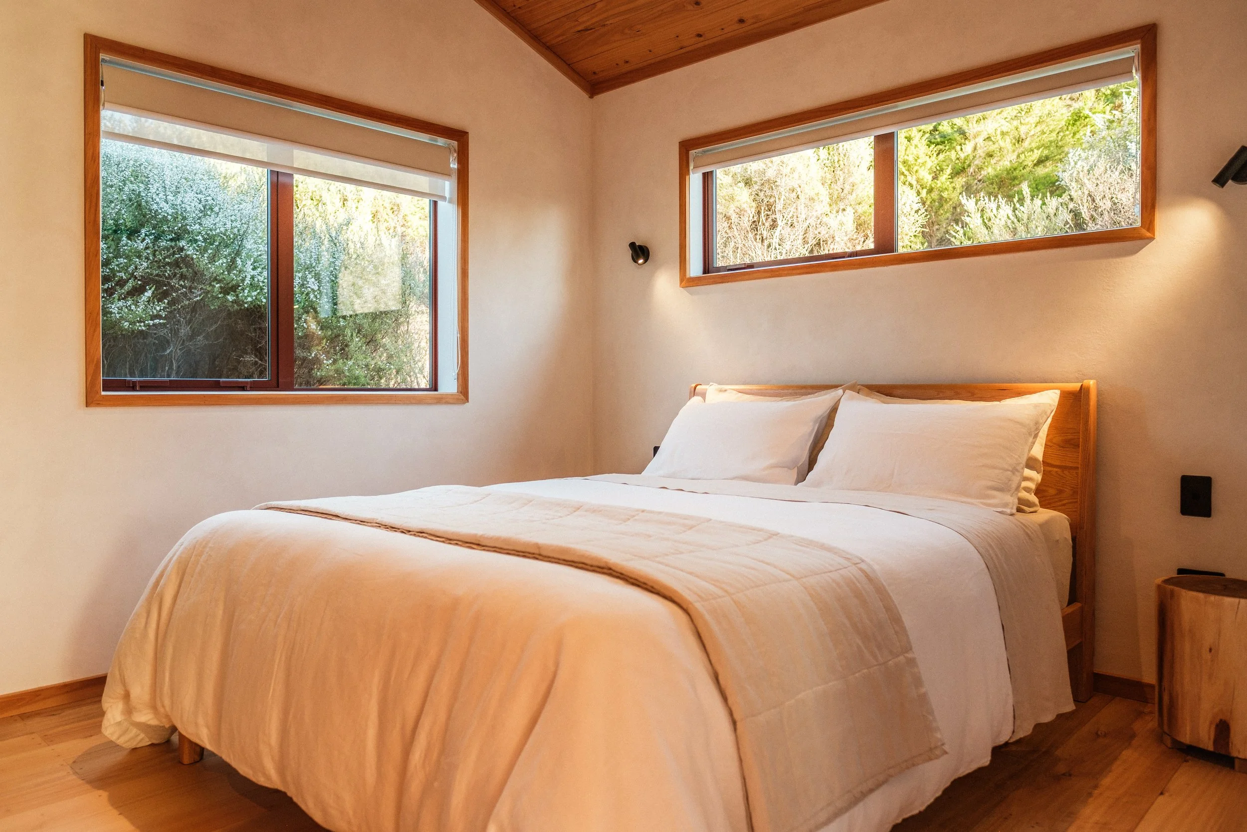 A cozy bedroom featuring a large bed with white bedding and pillows, a wooden headboard, and two windows showing greenery outside. The room has wooden accents on the ceiling, window frames, and a small side table, with soft lighting creating a warm a