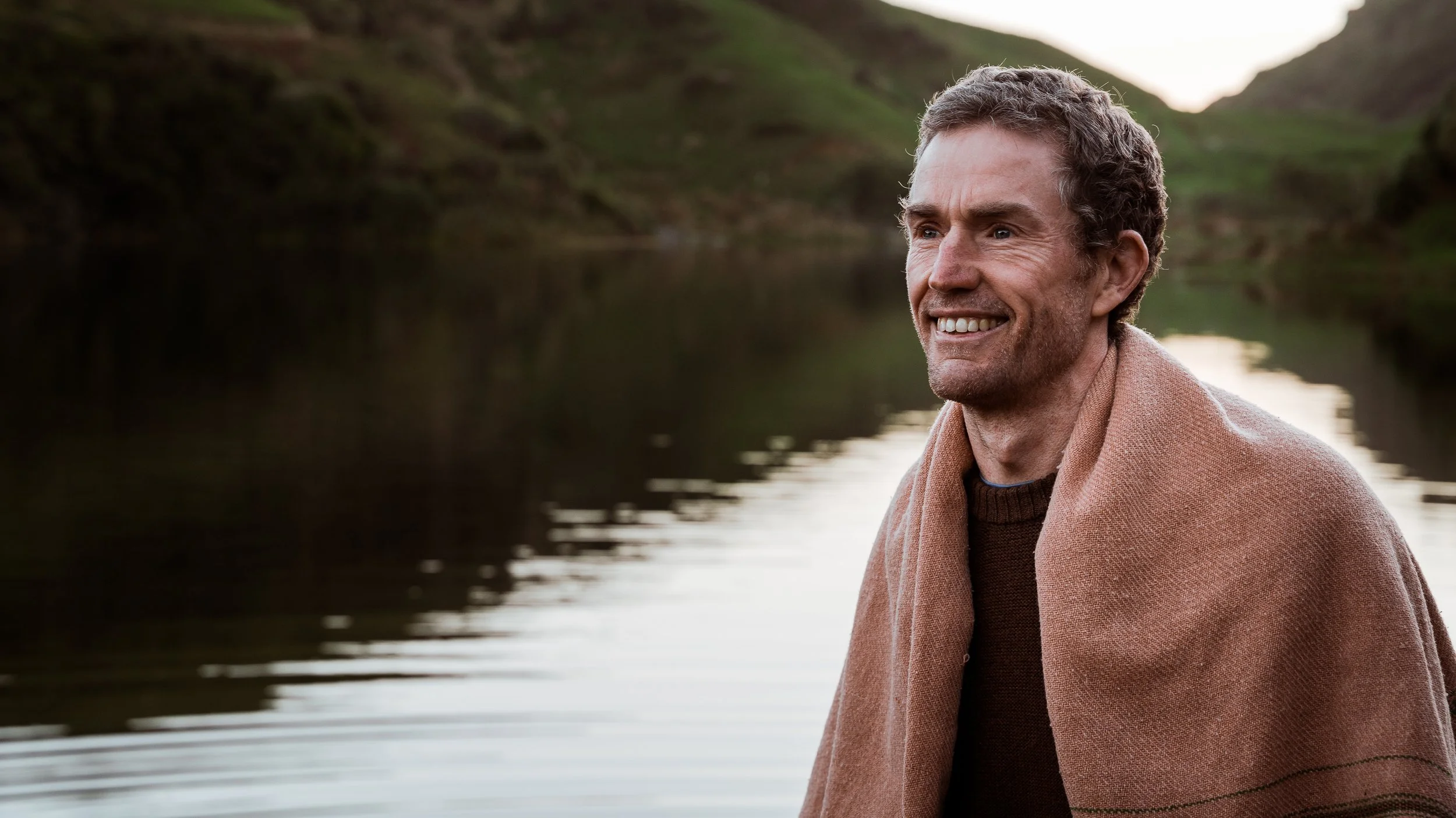 A man with curly hair and a beard smiling outdoors near a body of water, with green hills in the background, wearing a brown sweater and a tan shawl.