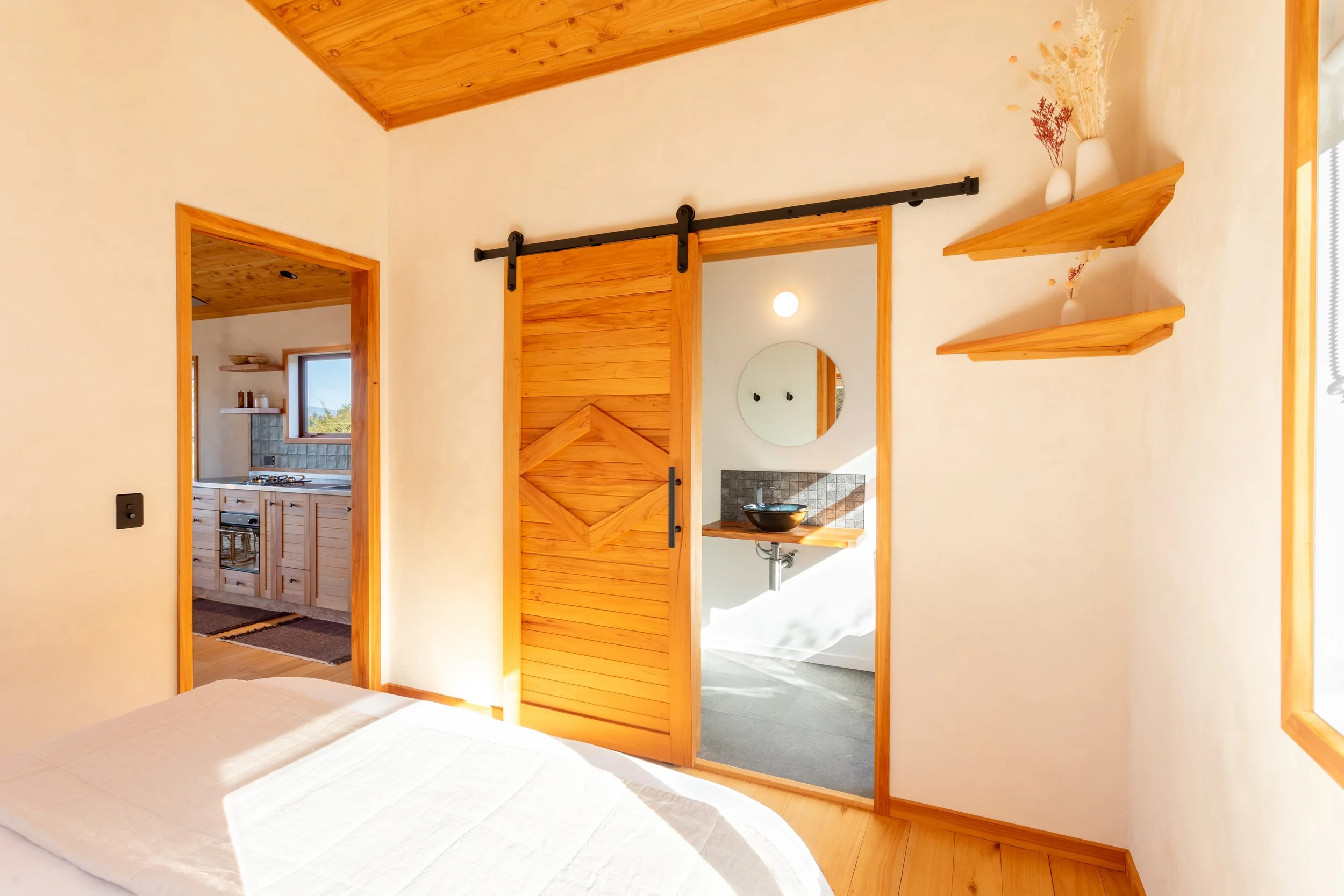 A bedroom with a wooden sliding barn door leading to an ensuite bathroom with a black vessel sink and mirror. Two wall-mounted shelves with vases and dried flowers are on the wall. Curtains are partially visible on the window at the right.