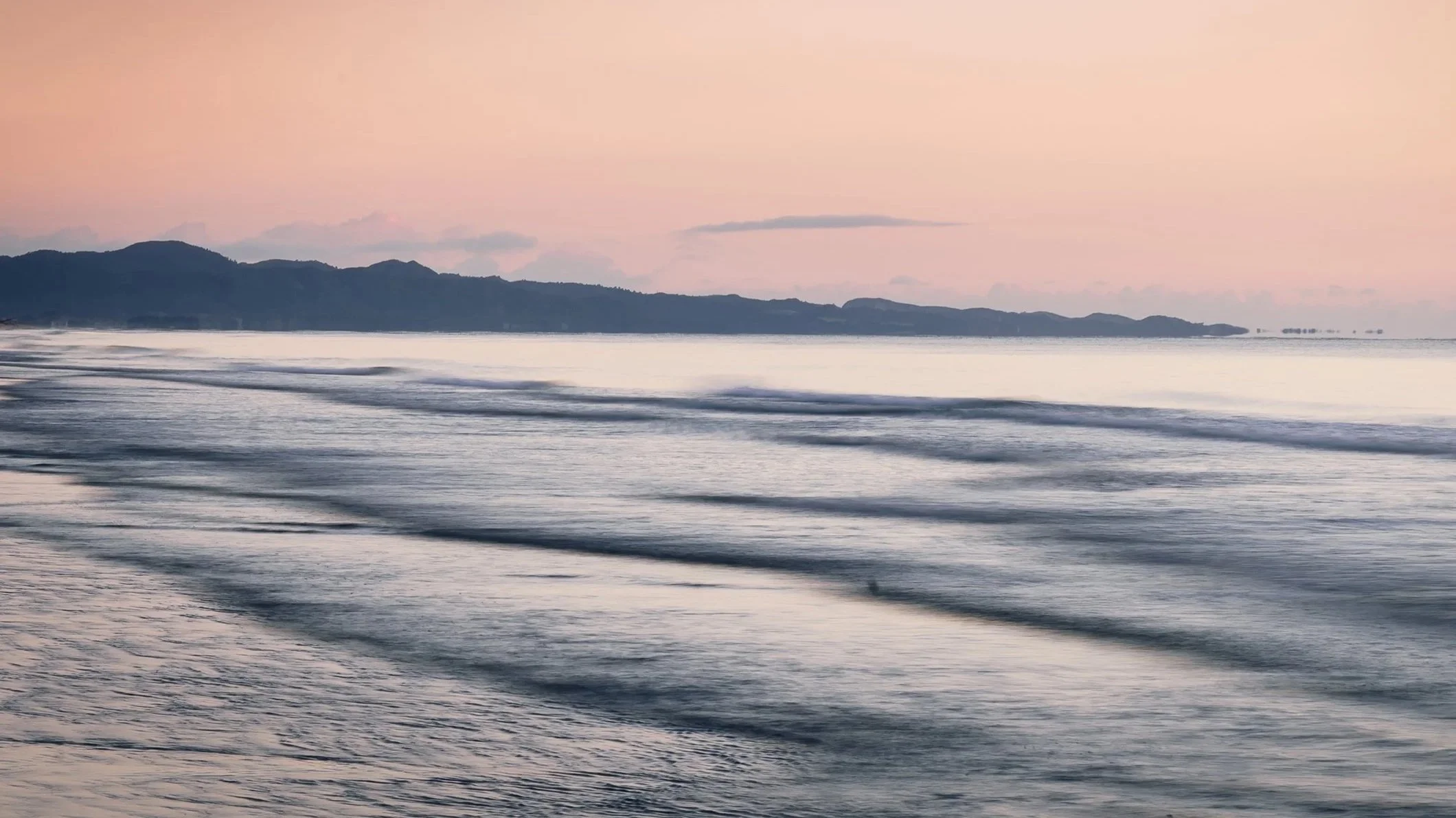 Calm ocean waves at sunset with a distant mountain range and pinkish sky.