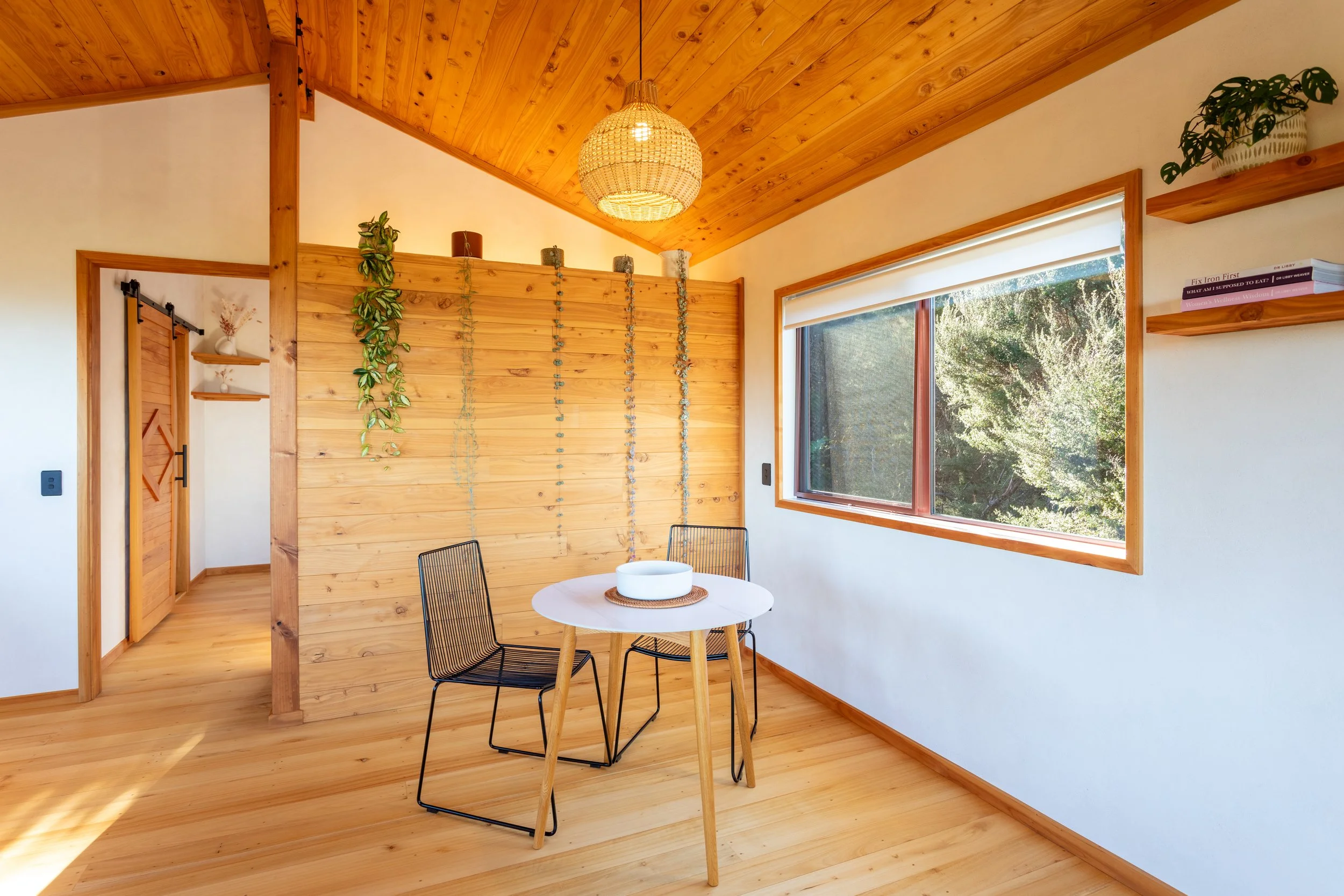 A cozy room with wooden walls, ceiling, and floor. There is a round dining table with two black wire chairs, a window with a white shade, and decorative plants and books on shelves and atop a wooden partition.