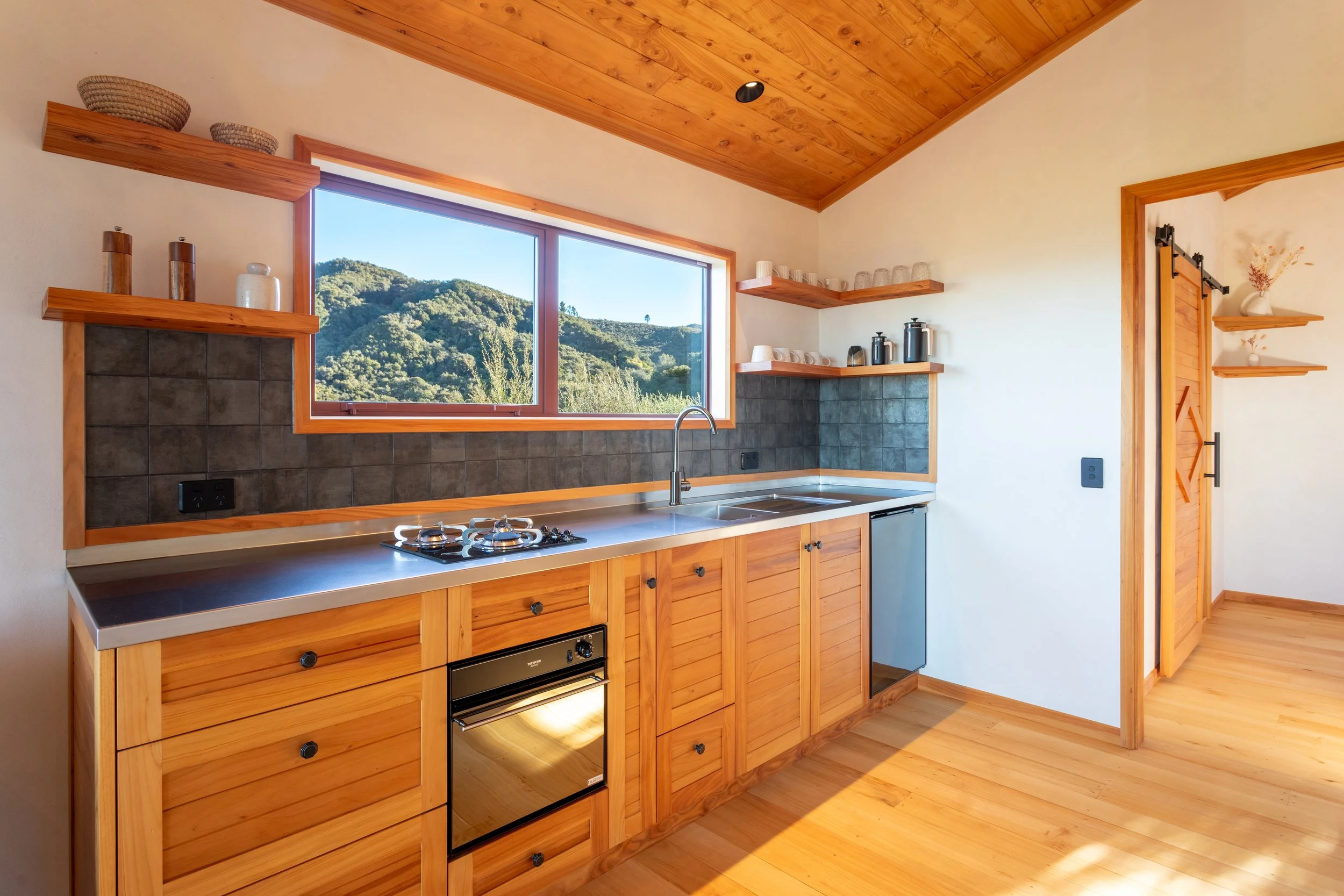 A kitchen with wooden cabinets and a countertop, a window showing a view of green hills, and shelves with dishes and kitchen appliances.