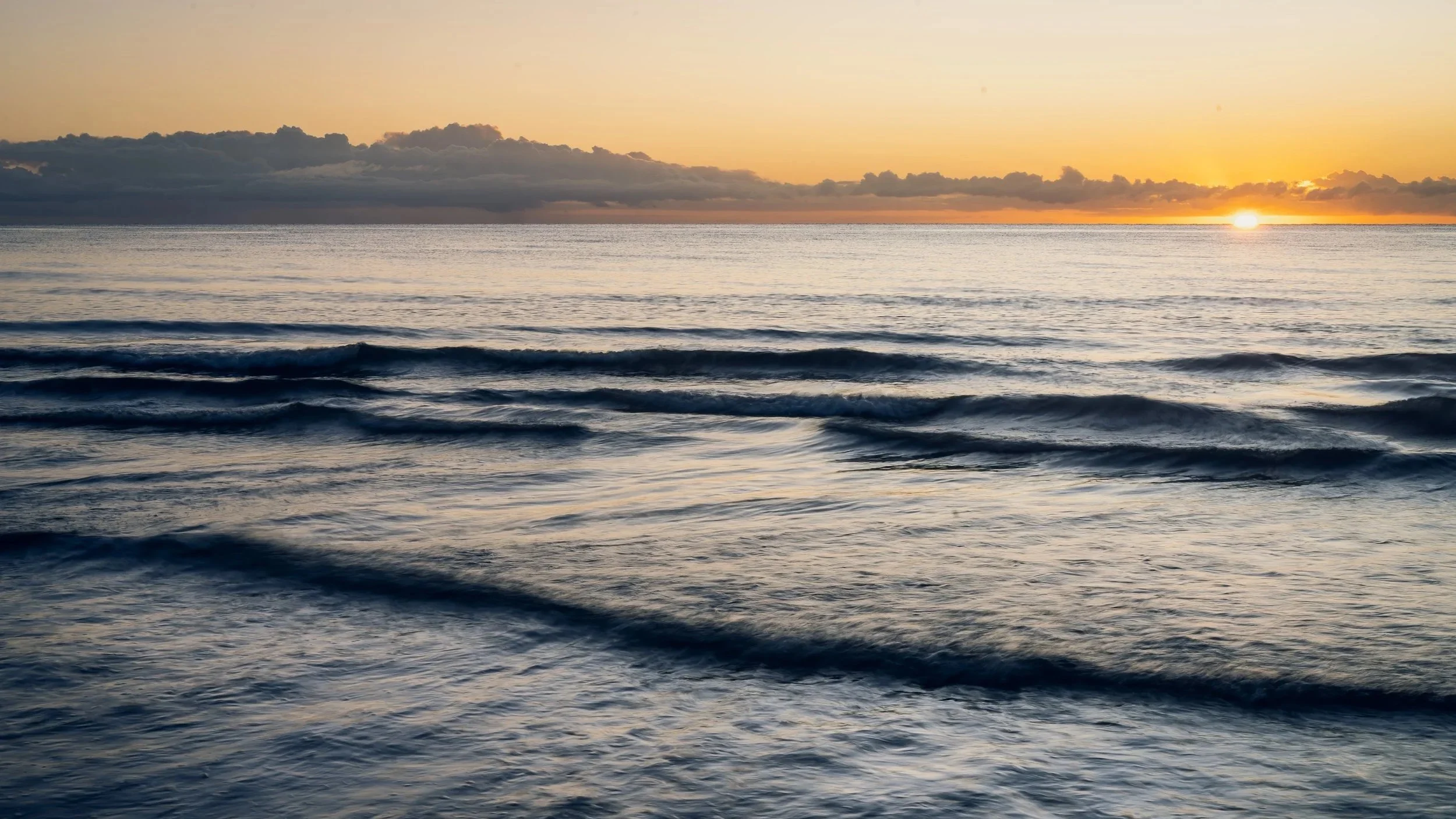 Sunset over the ocean with calm waves and dark clouds on the horizon.