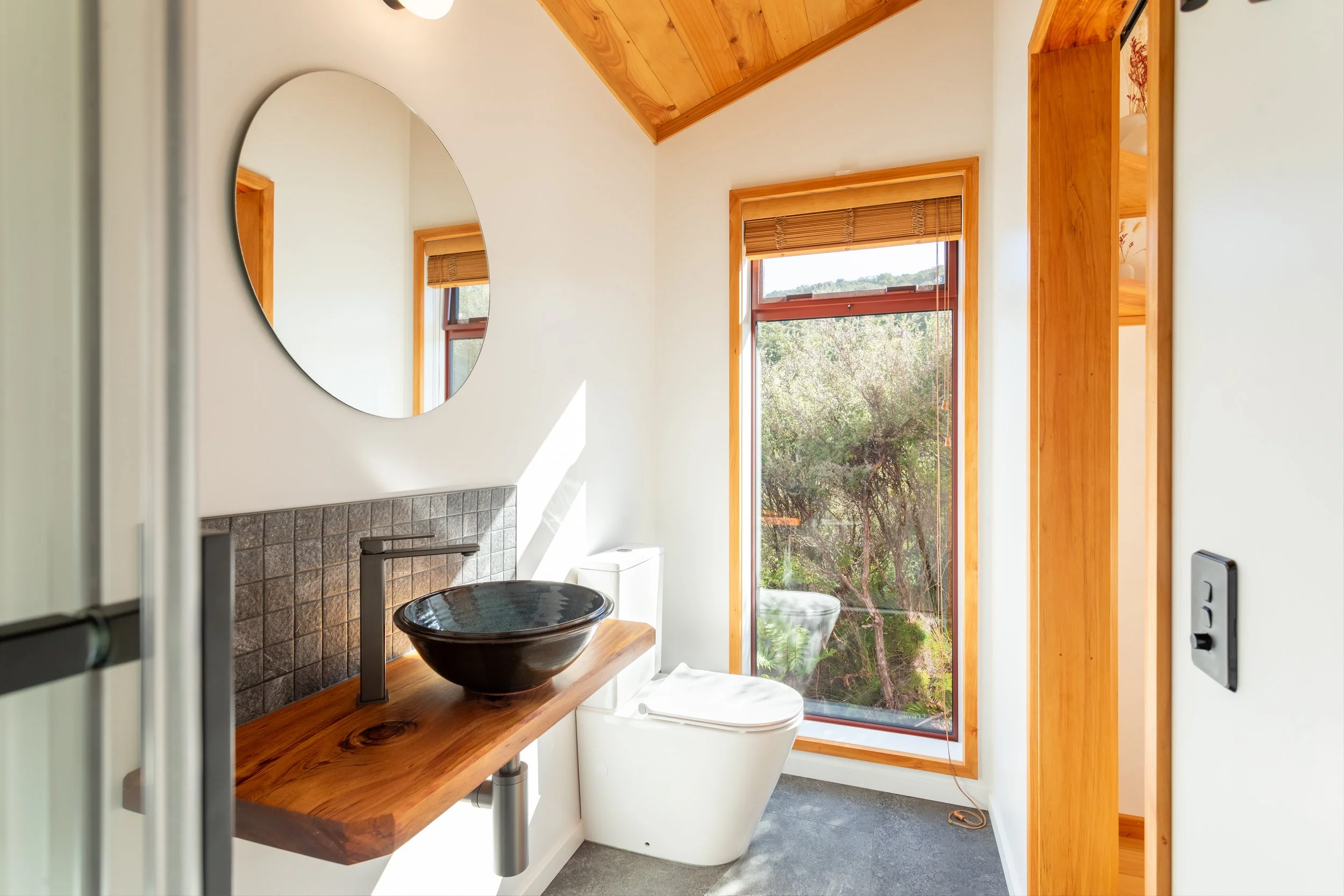 A small bathroom with a window overlooking trees, a wooden shelf with a black vessel sink, a modern toilet, and a large round mirror. The walls and ceiling are white, with wood trim around the window and ceiling.