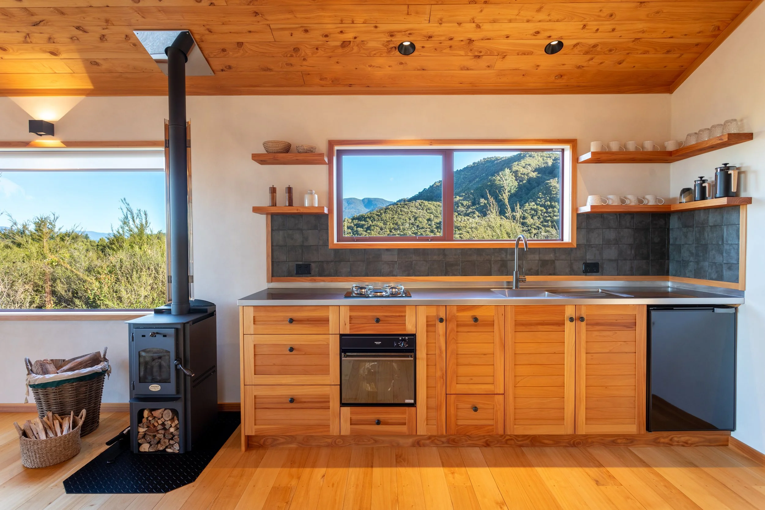 Kitchen with wooden cabinets, a large window showing a mountain view, a black wood stove, and open shelves with cups, bowls, and kitchen appliances.