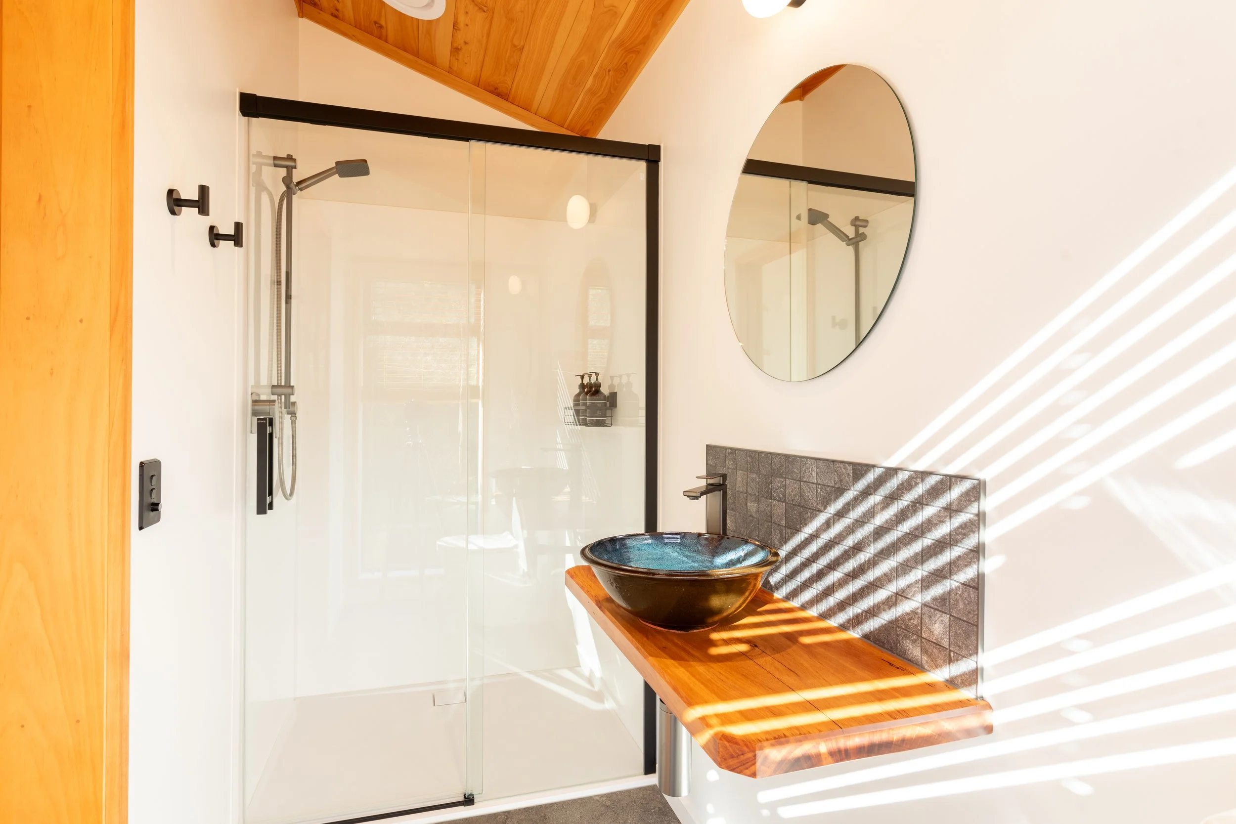 Modern bathroom with a glass shower enclosure, a round mirror on a white wall, a dark stone sink on a wooden countertop, and a textured dark tile wall behind the sink. There is natural sunlight creating shadows on the wall.