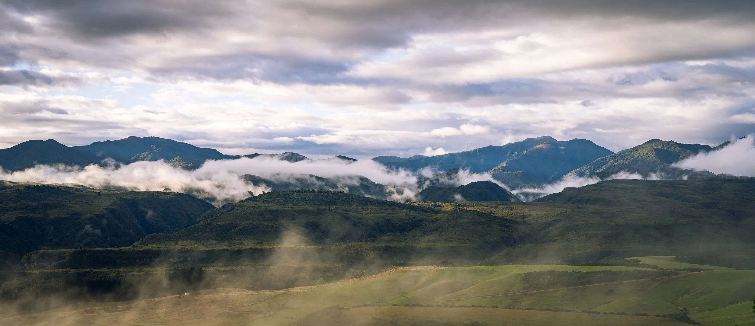 Mountain landscape with green hills, mist, clouds, and distant mountain peaks under a cloudy sky.
