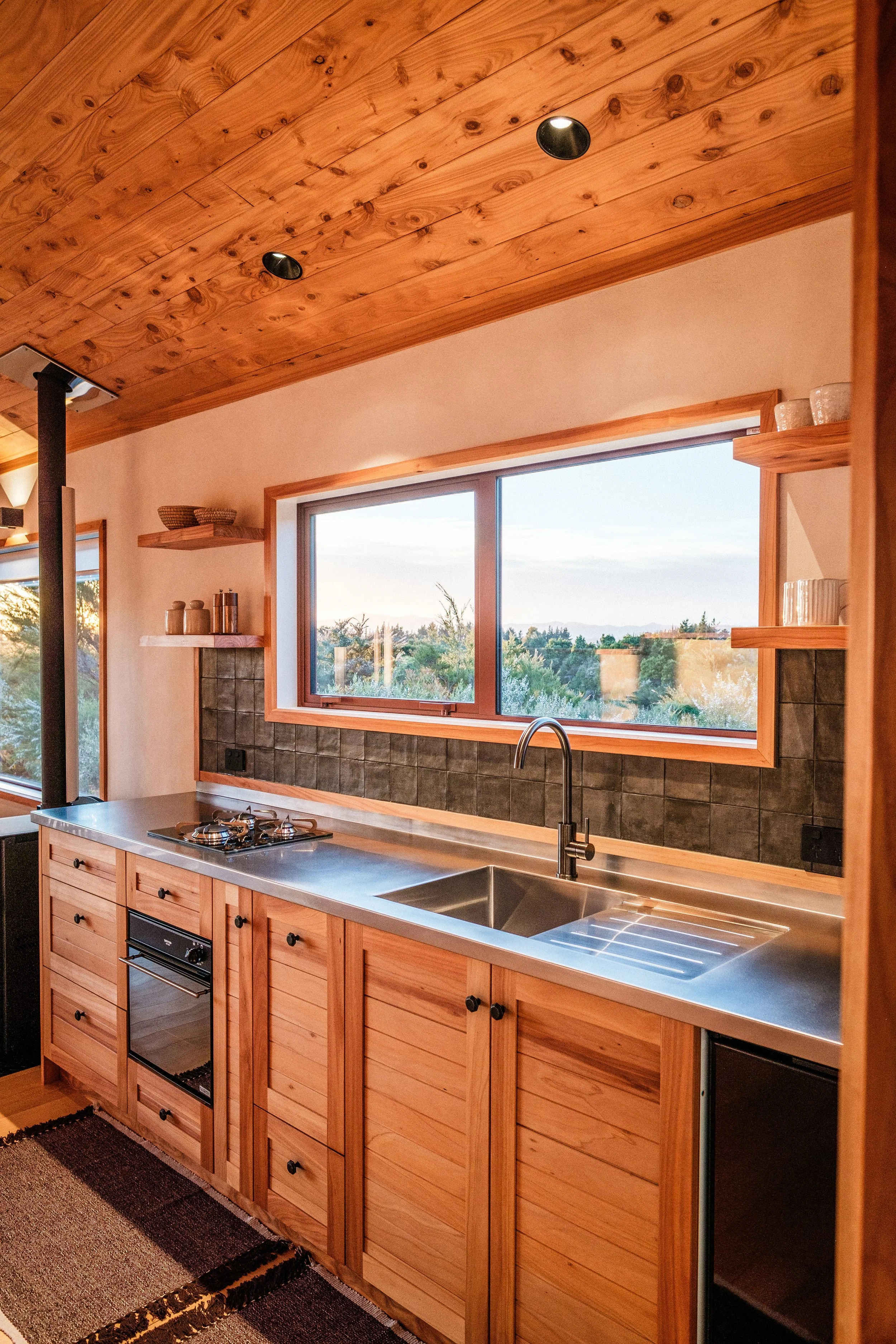 Cozy kitchen with wooden cabinets, stainless steel countertop, and a window overlooking a scenic landscape with trees; wood-panelled ceiling with premium lighting.