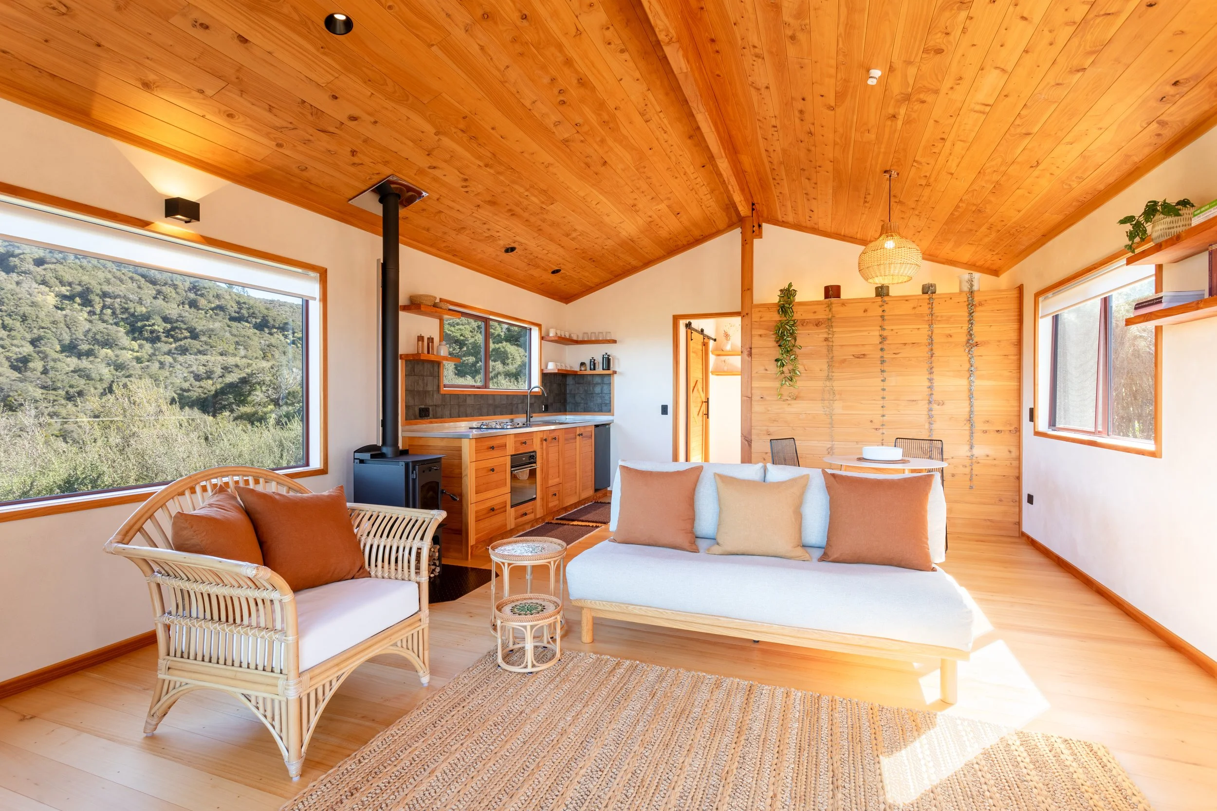 Living room with light wood walls and ceiling, large windows showing green landscape, white sofa with beige pillows, wicker chairs with beige cushions, small woven side table, kitchen area with wood cabinets, black stove pipe, and open shelves.