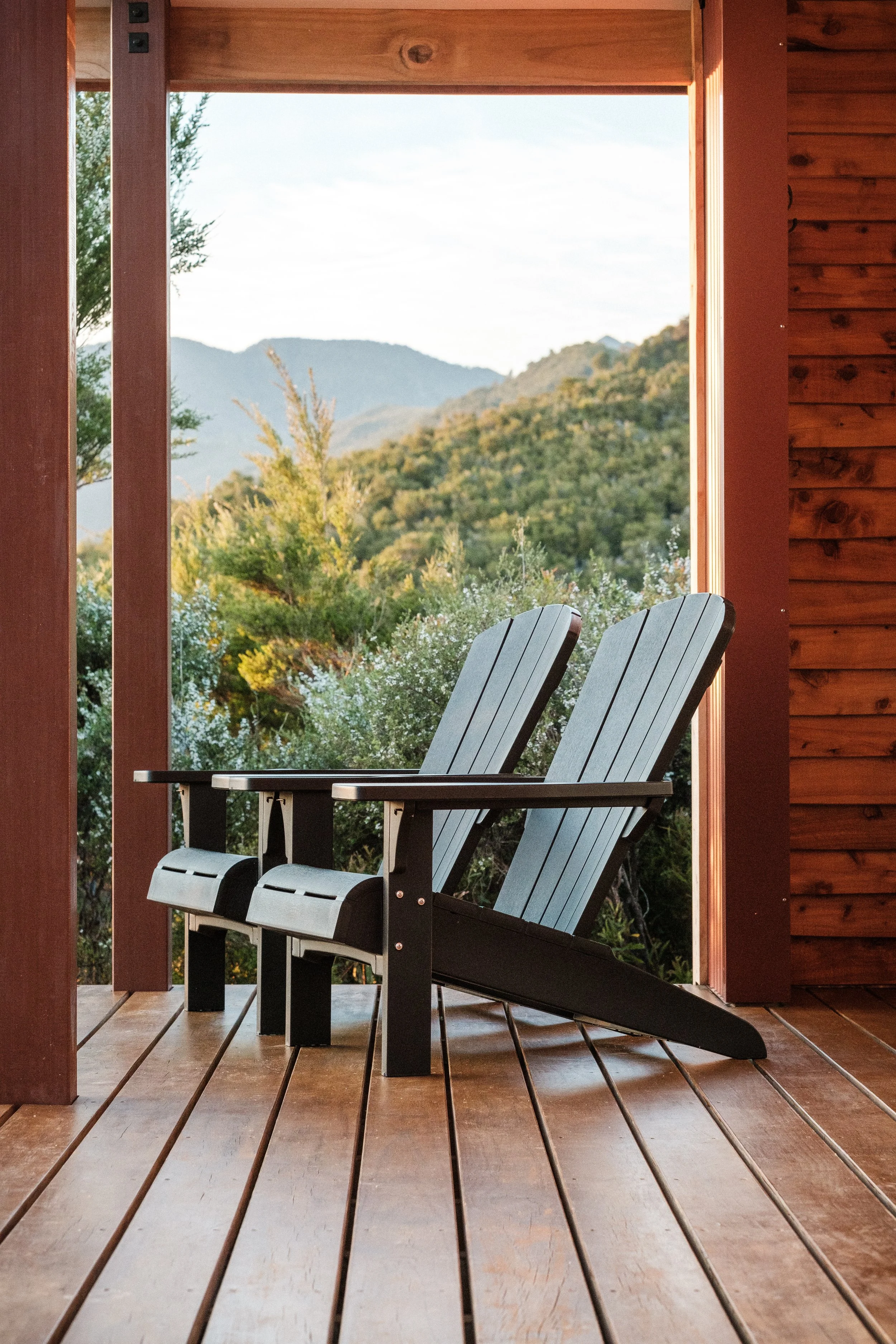 Two Adirondack chairs on a wooden porch overlooking a lush green mountainous landscape with trees and hills in the distance.