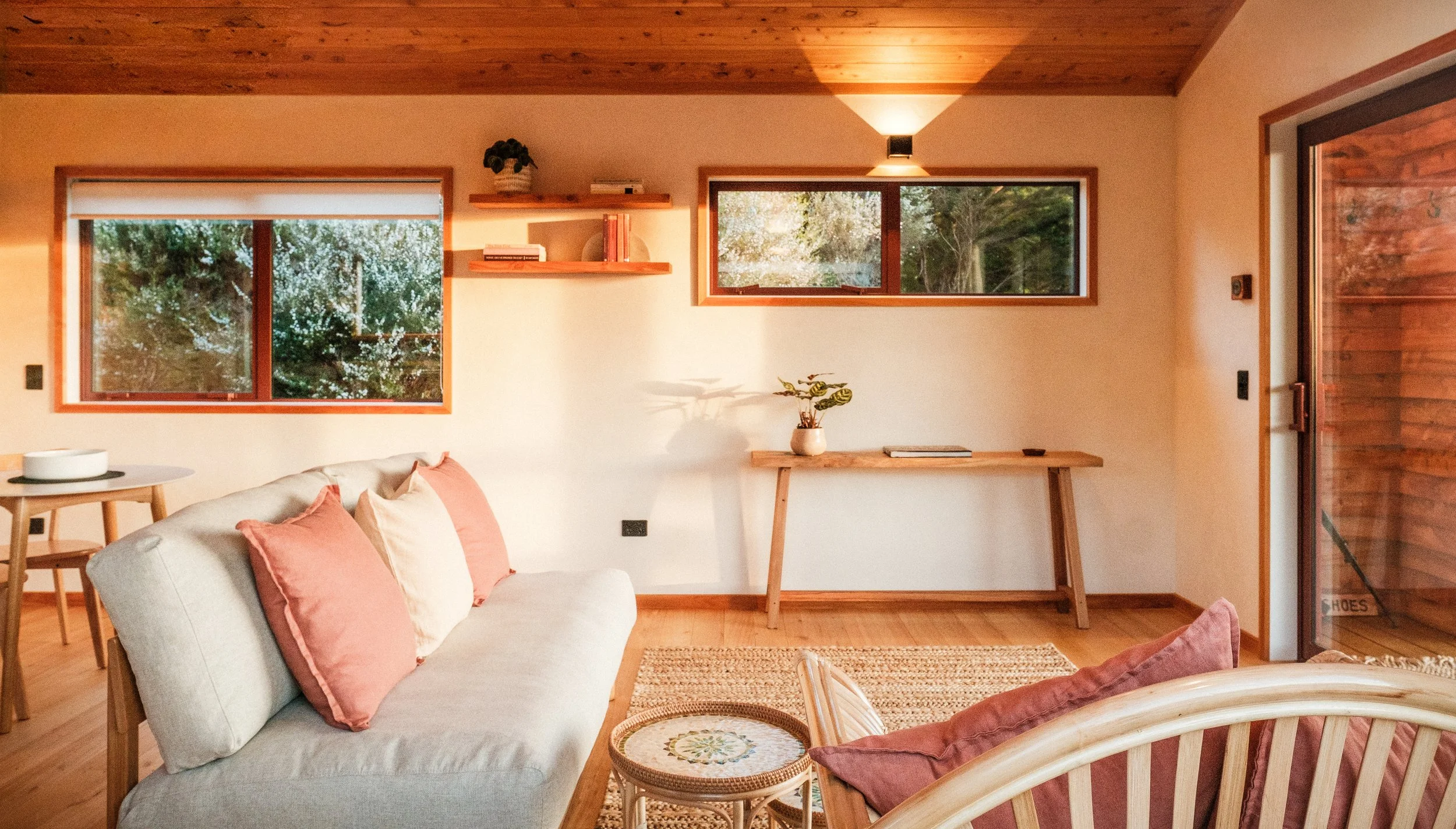 Living room with a beige sofa with pink pillows, a small round table, a wooden desk, and windows showing outdoor greenery.