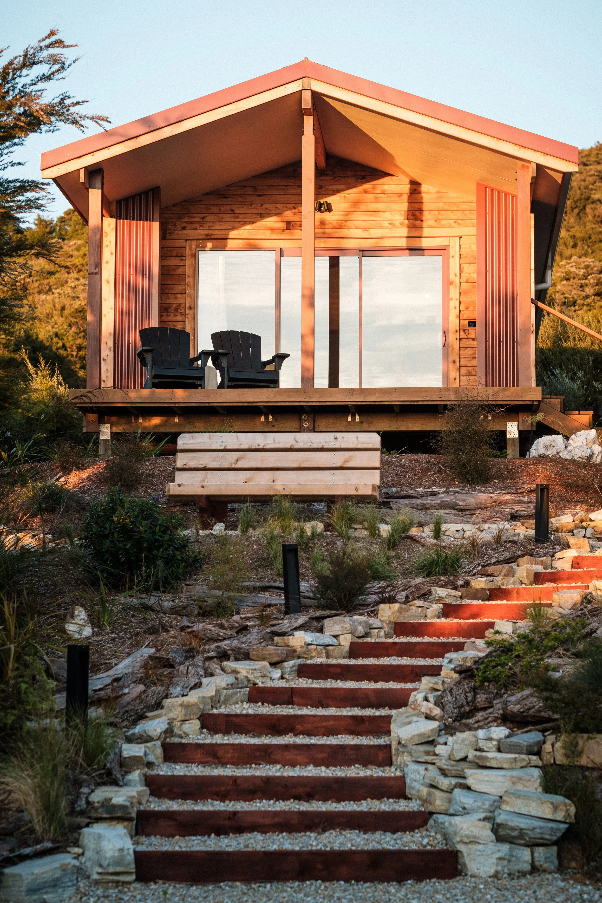 A private nature retreat with a red roof on a hillside, accessed by a stepped stone and wooden staircase, with two black chairs on the porch, during sunset.