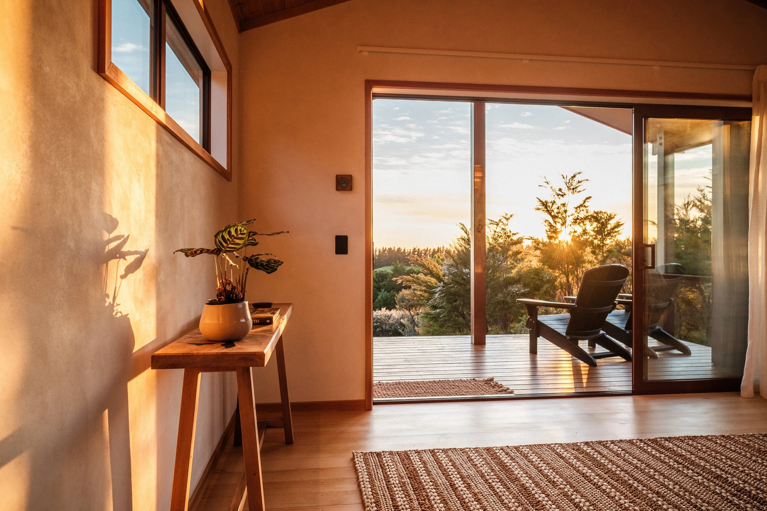 Interior of a room with a sliding glass door opening to a deck with a lounge chair, trees, and sunset view