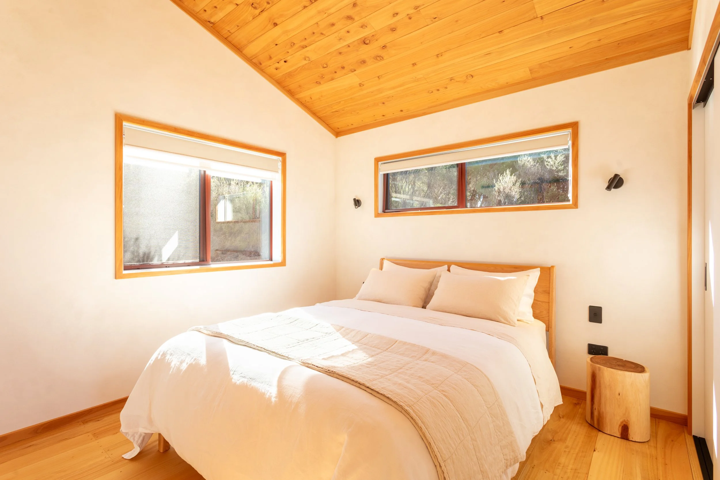 A cozy bedroom with a wooden ceiling, beige walls, and two windows letting in natural light. The bed has white bedding and a beige quilt, with a small round wooden side table beside it. The room has modern black wall-mounted reading lights.