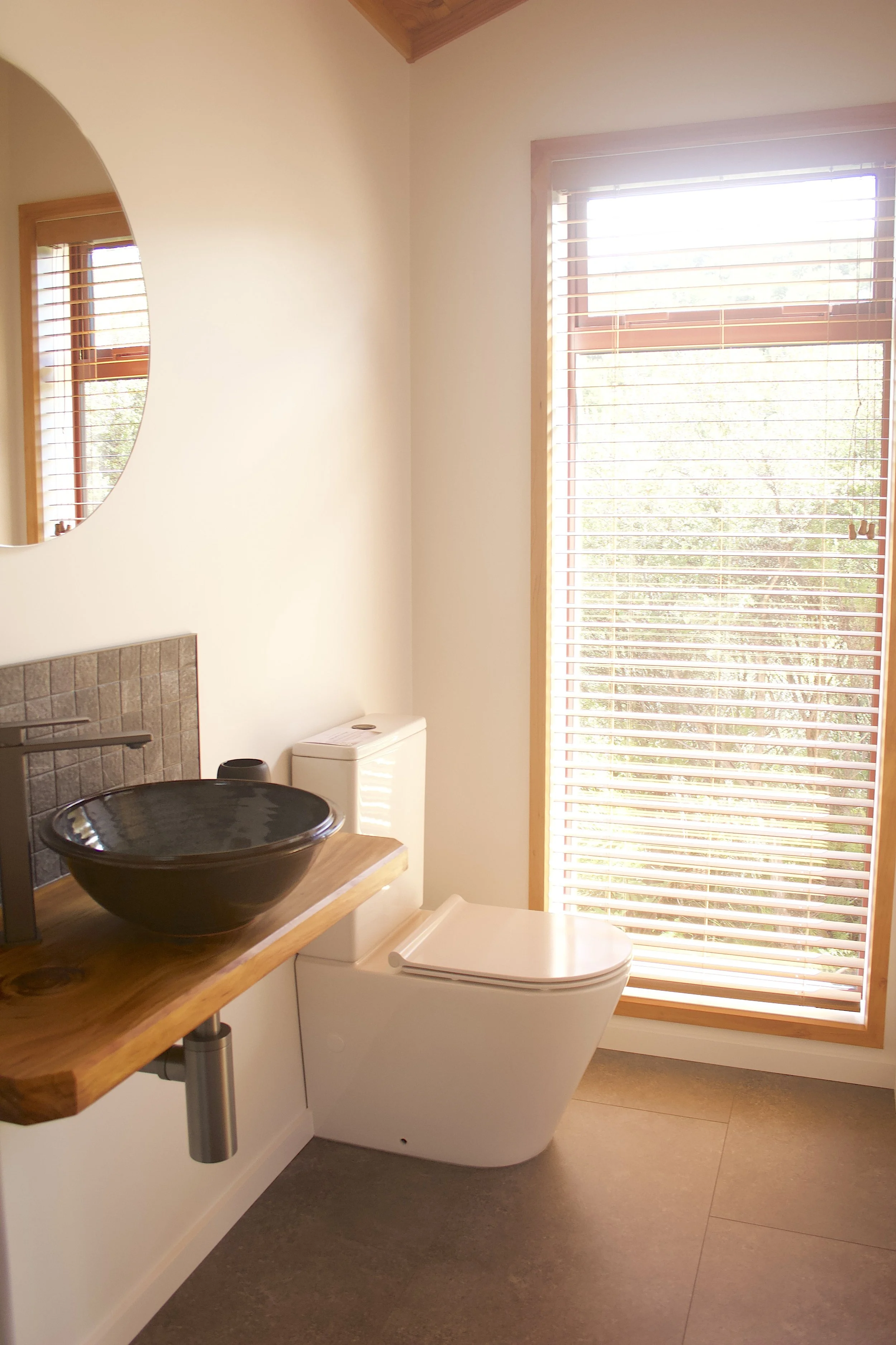 A minimalist bathroom with a black ceramic sink on a wooden countertop, a white toilet, a large window with wooden blinds, a round mirror, and neutral-coloured walls and flooring.
