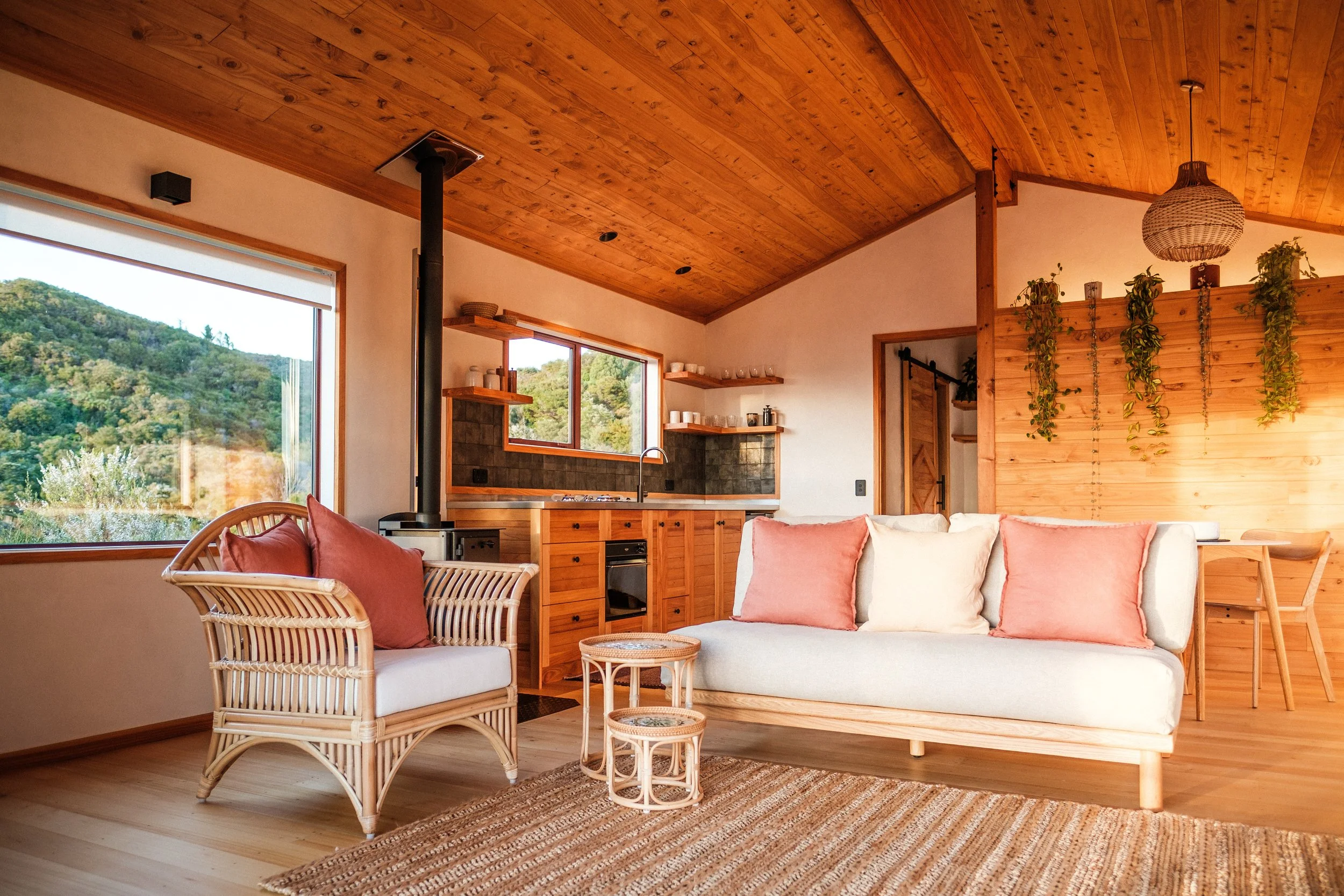 Living room with rattan furniture, wooden ceiling and walls, large windows showing green hills, and a cozy, modern style sofa and armchair with cushions. Plants hanging from a wooden wall.