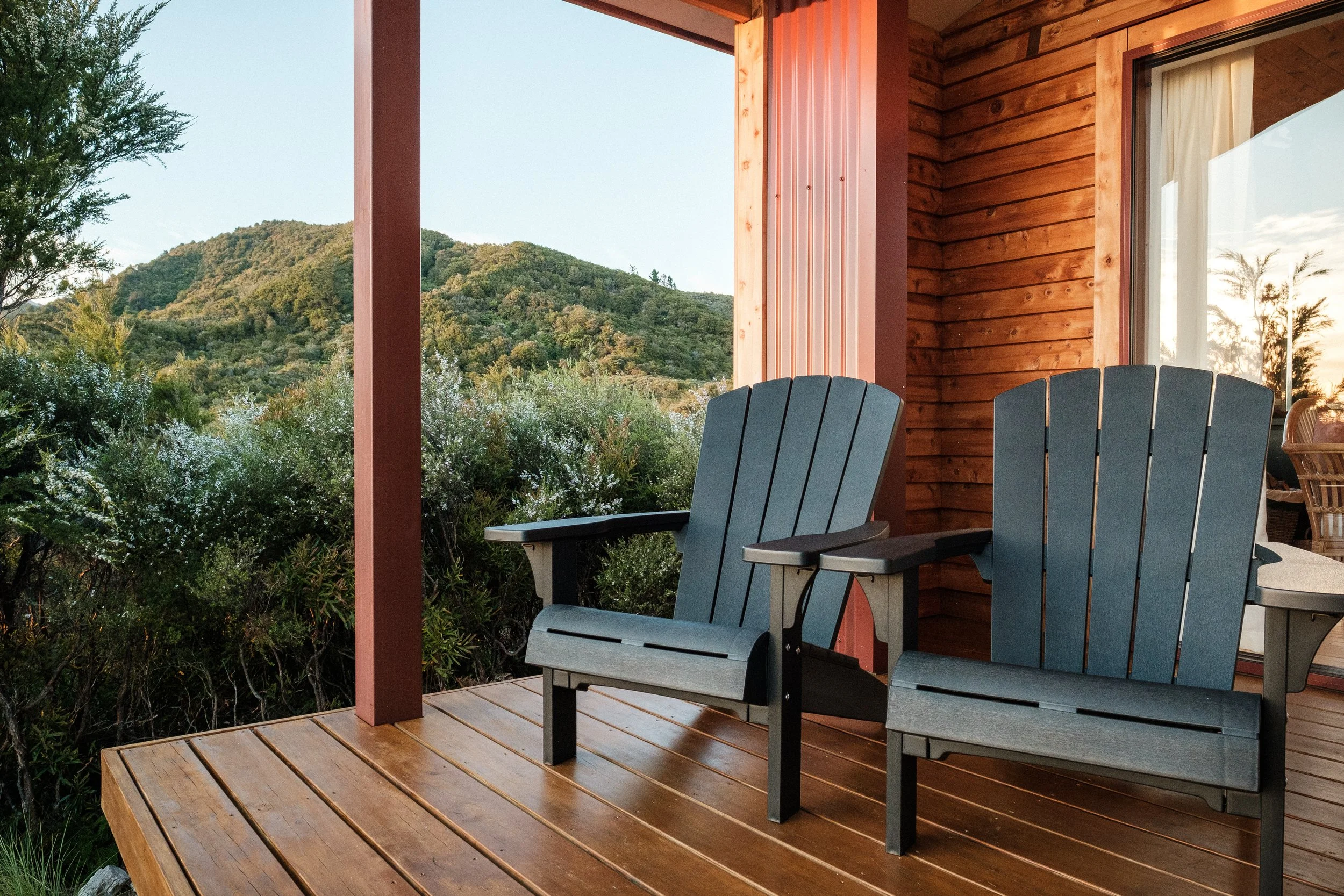 Two dark gray Adirondack chairs on a wooden porch overlooking lush green hills and trees at sunset.