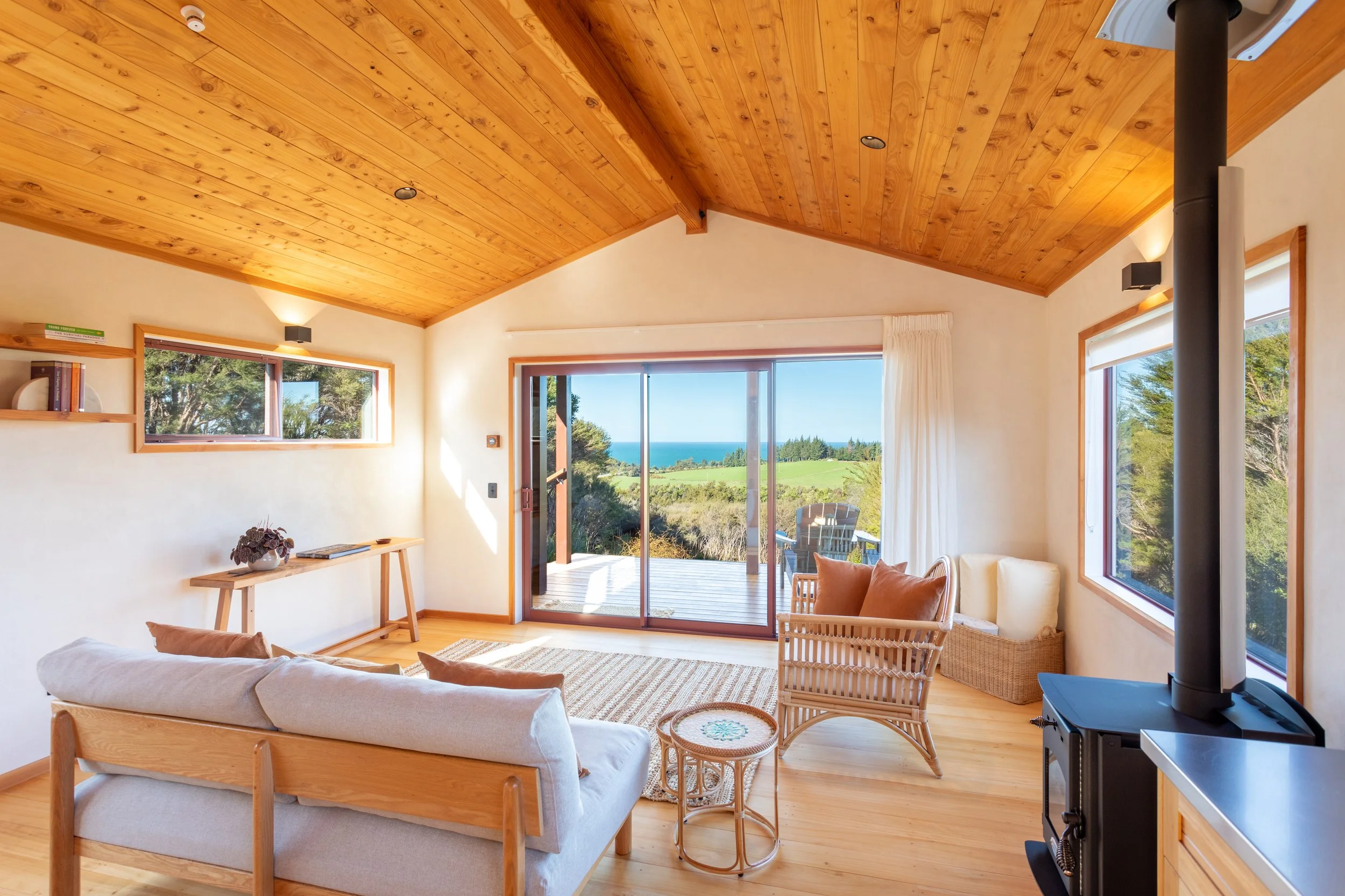 Living room with wooden ceiling and large windows; view of greenery and ocean outside; beige couch, rattan chair, side table with decorative pillow, heater pipe, and built-in shelf with books.