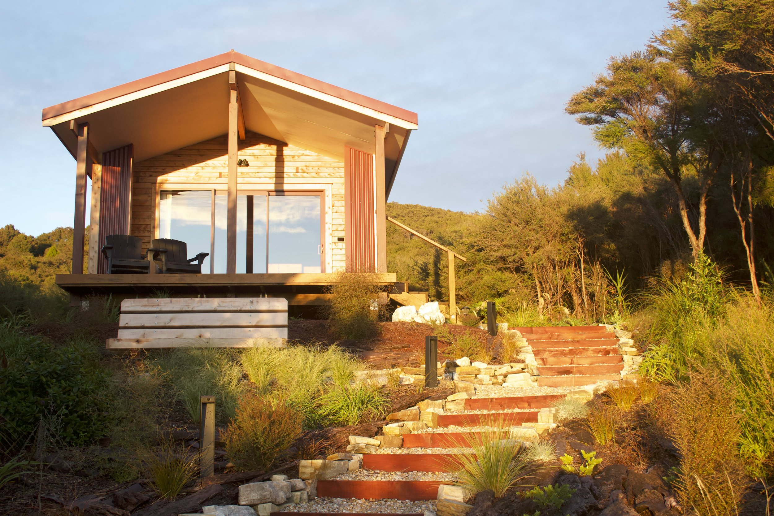 A small wooden house with a sloped roof, large windows, and a balcony with two chairs, situated on a hill surrounded by trees and greenery, with stone stairs leading up to it and the sky in the background.