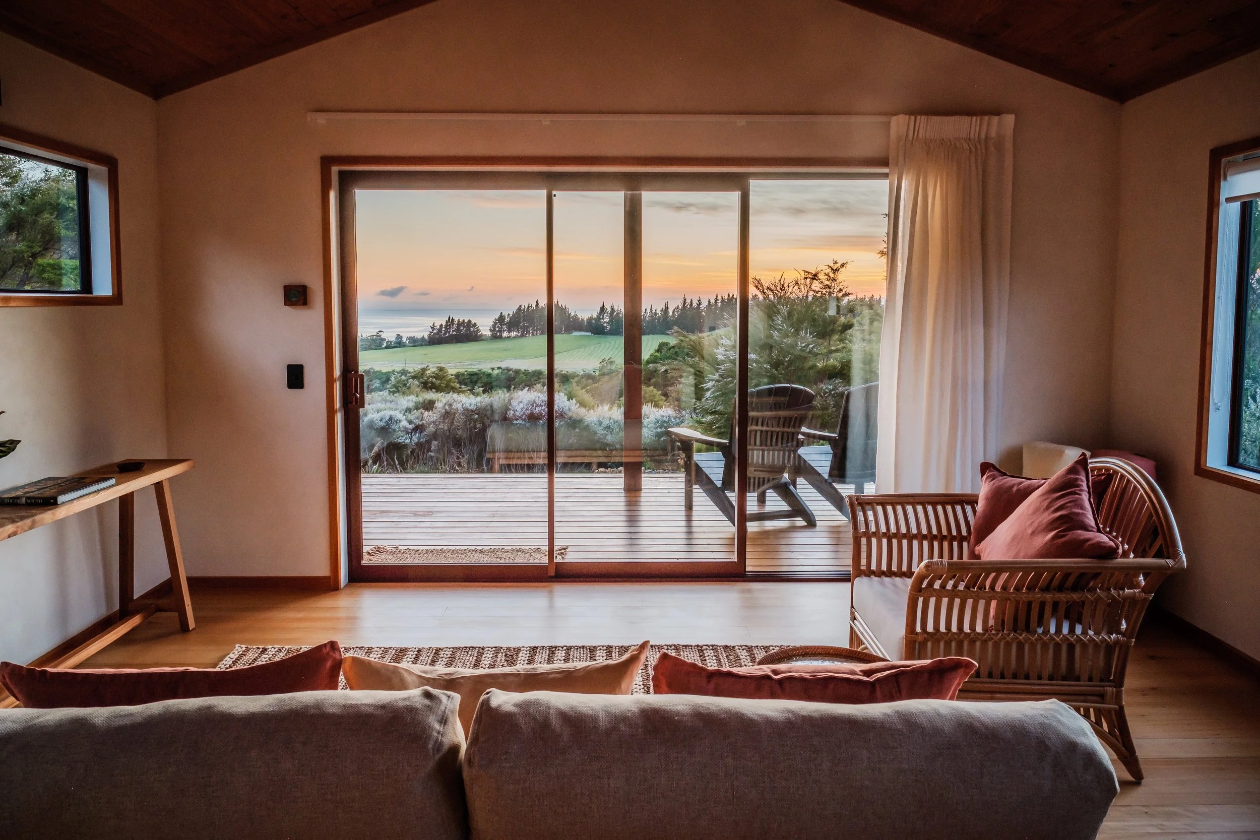 Living room with a large glass sliding door opening to a deck with outdoor chairs, overlooking a scenic landscape at sunset with trees and open fields.