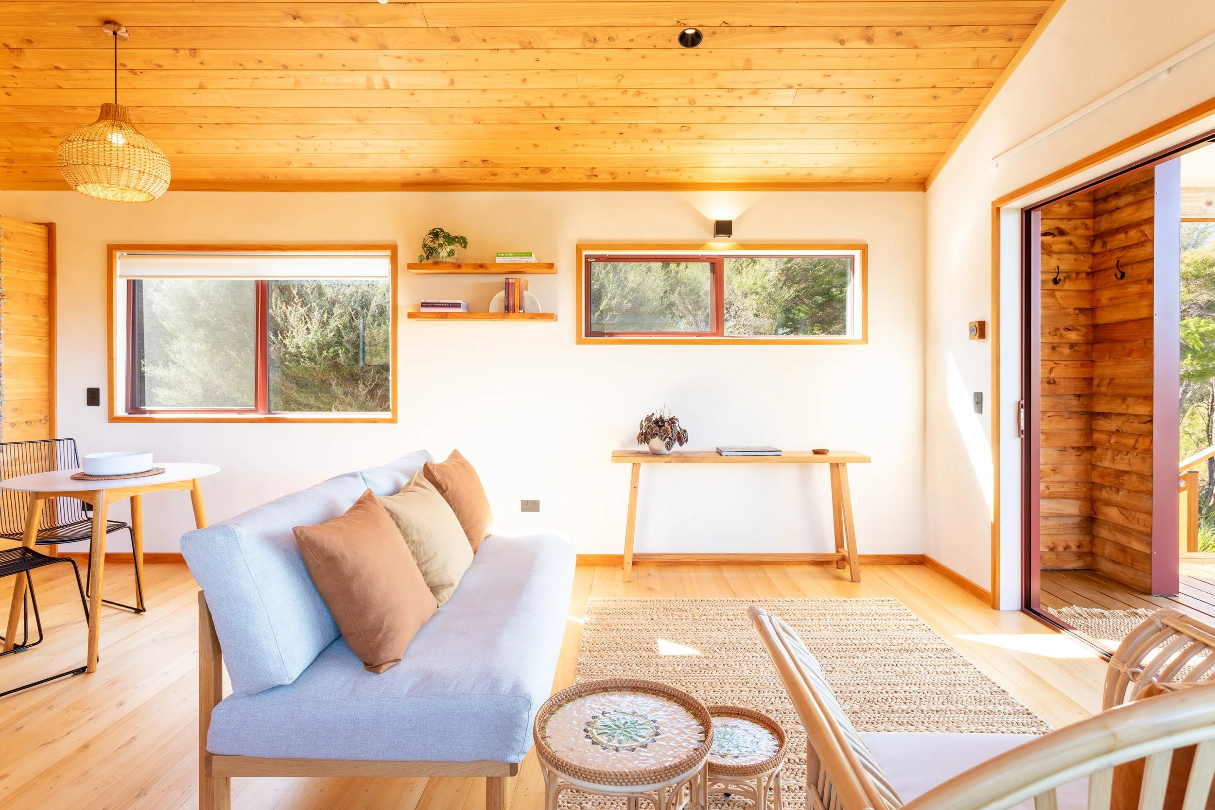 Living room with wooden ceiling and floors, cream walls, large windows showing greenery outside, beige sofa with brown pillows, small dining table with black chair, shelves with books and plants, woven light fixture, and patio visible through open do