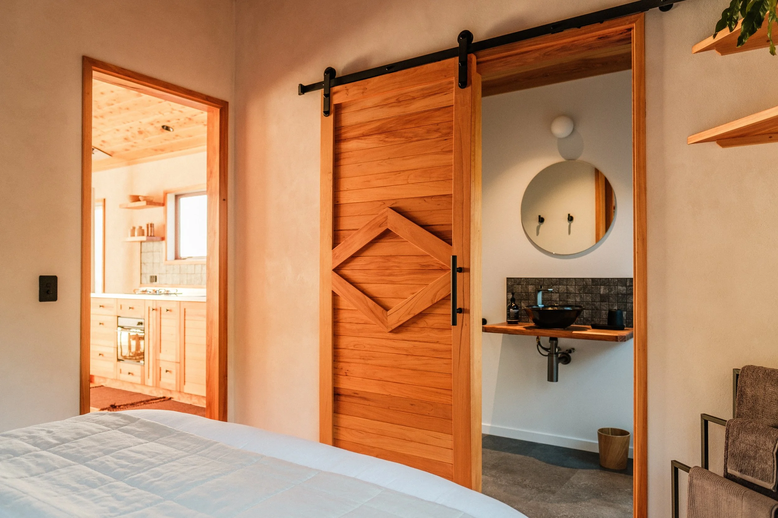 View of a bedroom with a wooden sliding door leading to a bathroom, and part of a kitchen visible through an open doorway, in a cozy, rustic home.