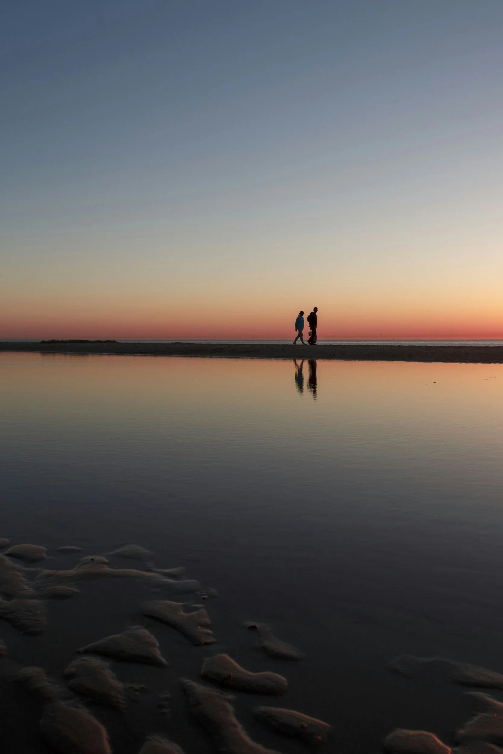 Silhouetted group of three people walking along a beach at sunset, with their reflections visible in the water, and smooth rocks in the foreground.