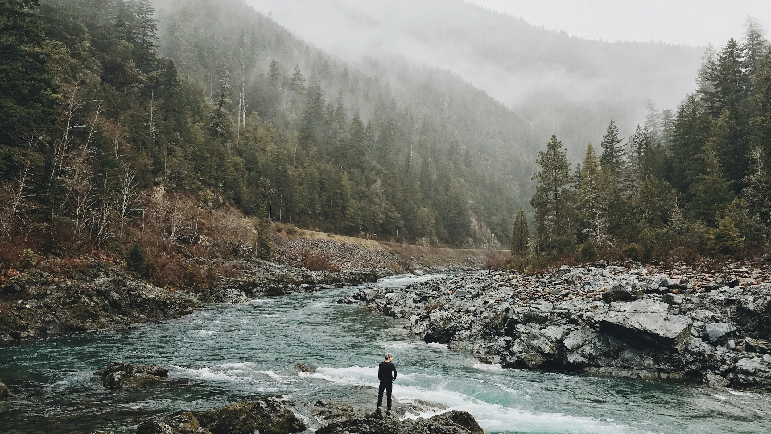 A person standing on rocks by a rushing river in a mountainous forest landscape with misty trees in the background.