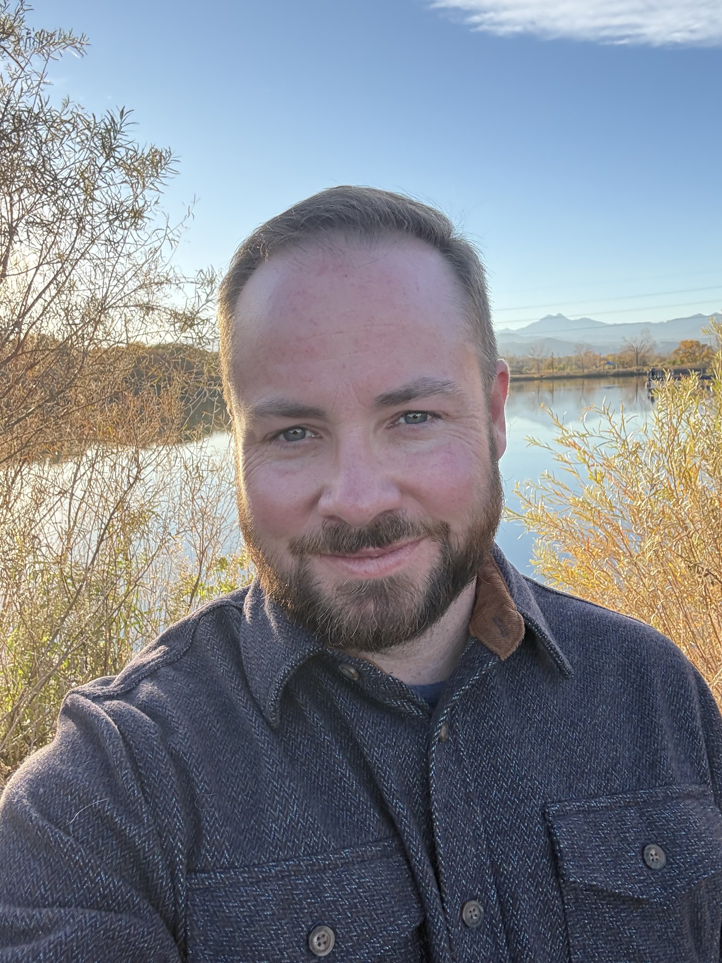 A man with a beard and short hair smiling outdoors during sunset near a lake with trees and mountains in the background.