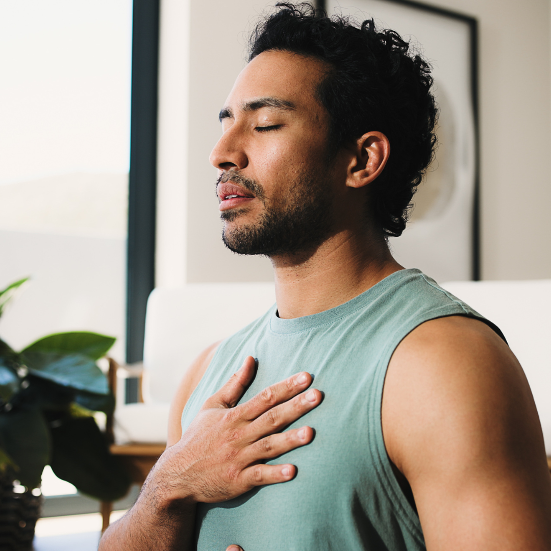 Man with dark curly hair and a beard practicing mindfulness with hand on chest, eyes closed, indoors near window.