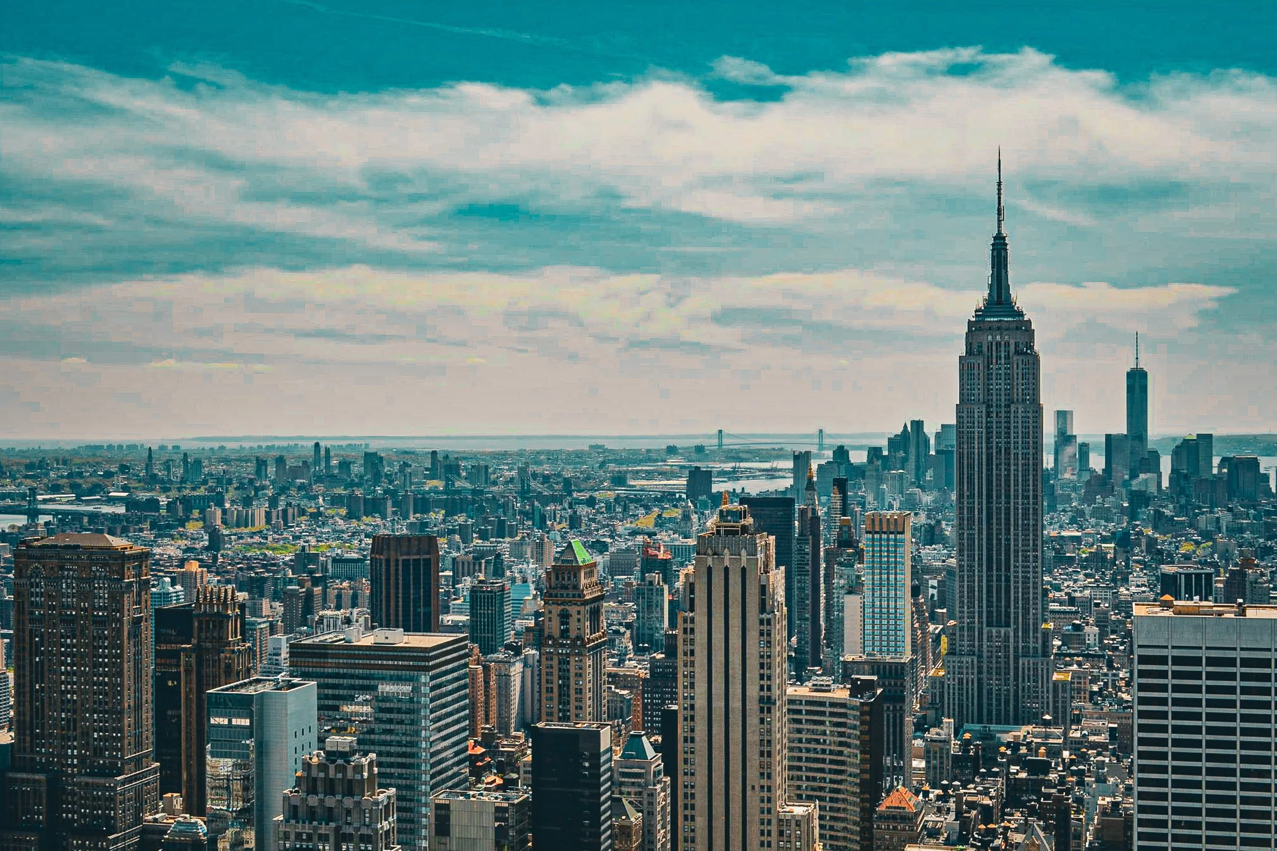 The Empire State Grid - A wide-angle cityscape photograph of the New York City skyline, highlighting the sharp architectural geometry of the Empire State Building towering over a dense grid of skyscrapers under a cloudy sky.