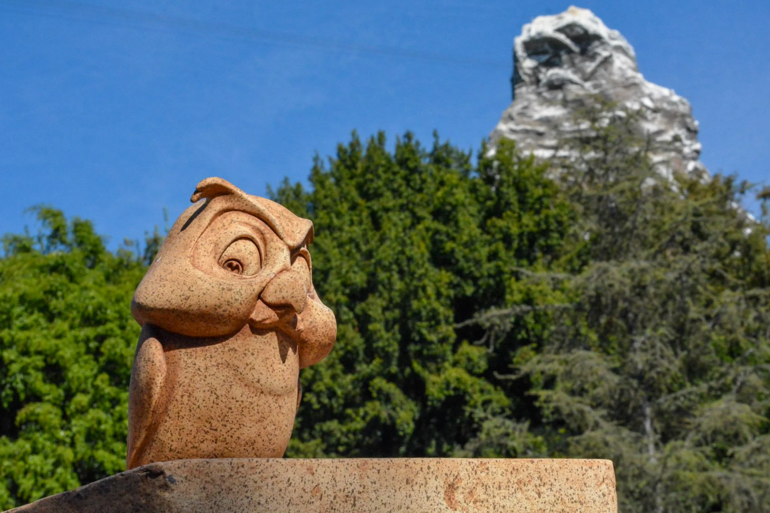 Mountain View Owl - A delightful companion shot featuring the same expressive stone owl statue, positioned on the left to frame a snow-capped, rugged mountain peak rising above a dense green treeline in the distance.