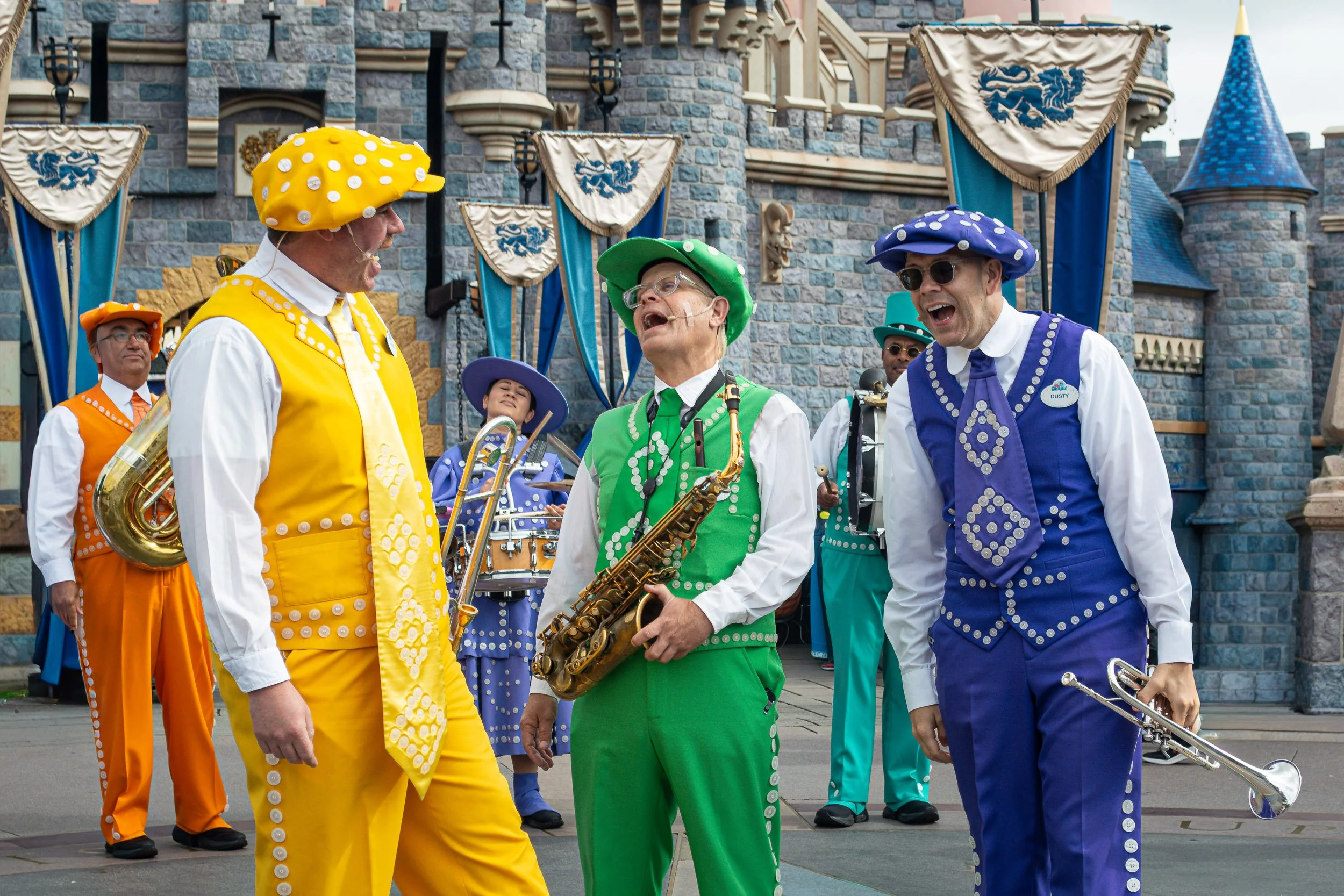 Dapper Harmony - A candid, high-energy shot of the Dapper Dans singing group performing in vibrant, custom-buttoned suits of yellow, green, and purple in front of a stone castle facade.