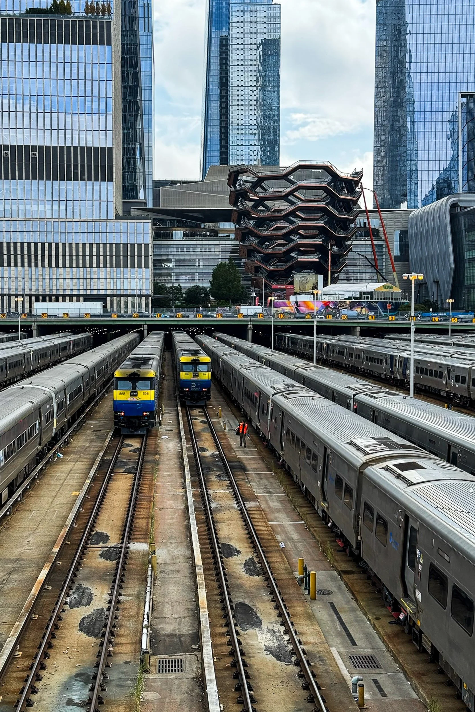 High-angle view of a city rail yard with silver and blue trains on parallel tracks leading toward glass skyscrapers and the bronze geometry of The Vessel.