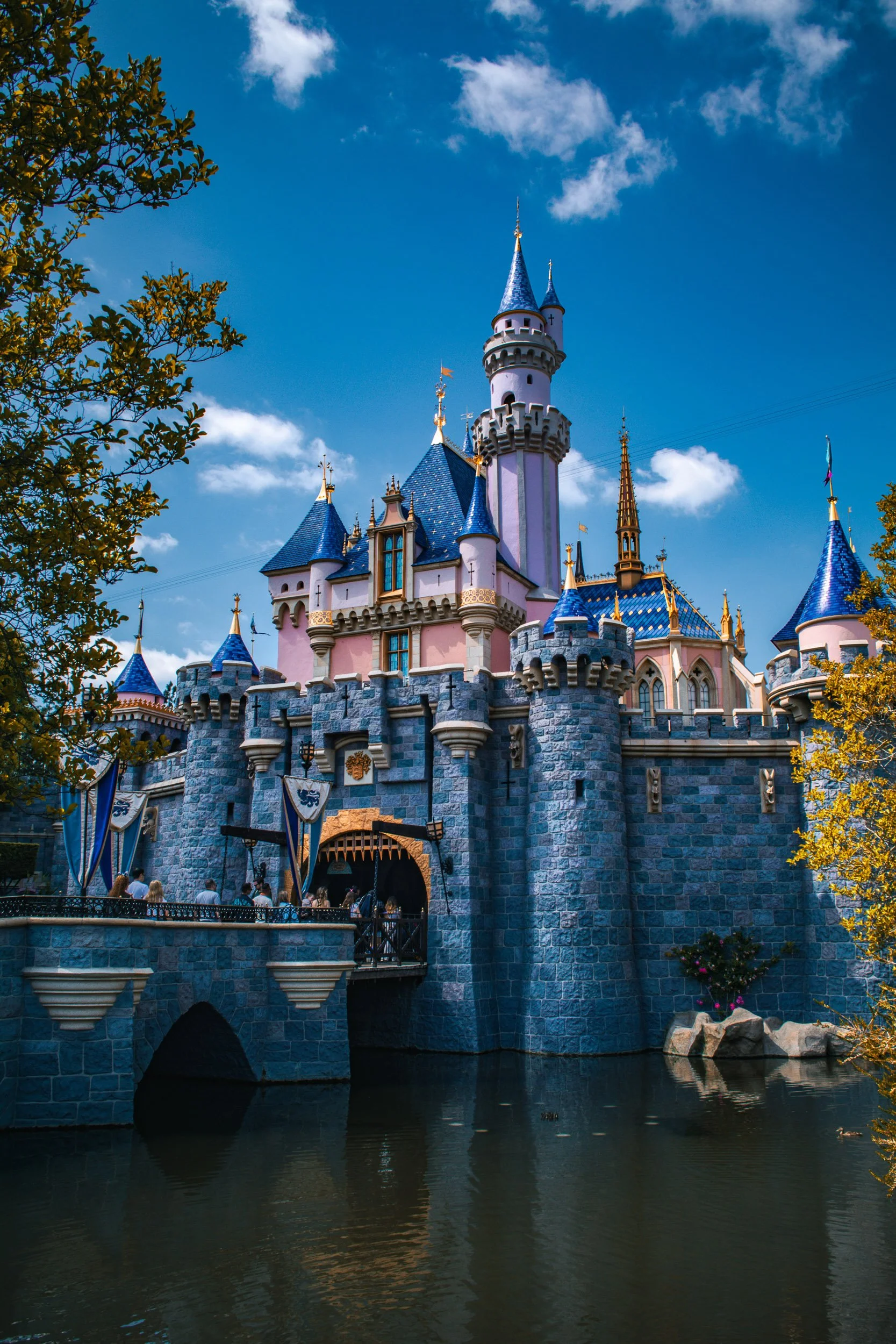 Fairytale Spire - A majestic vertical perspective of Sleeping Beauty Castle, highlighting its pink walls and brilliant blue turrets reaching into a crisp sky framed by rich, sunlit foliage.