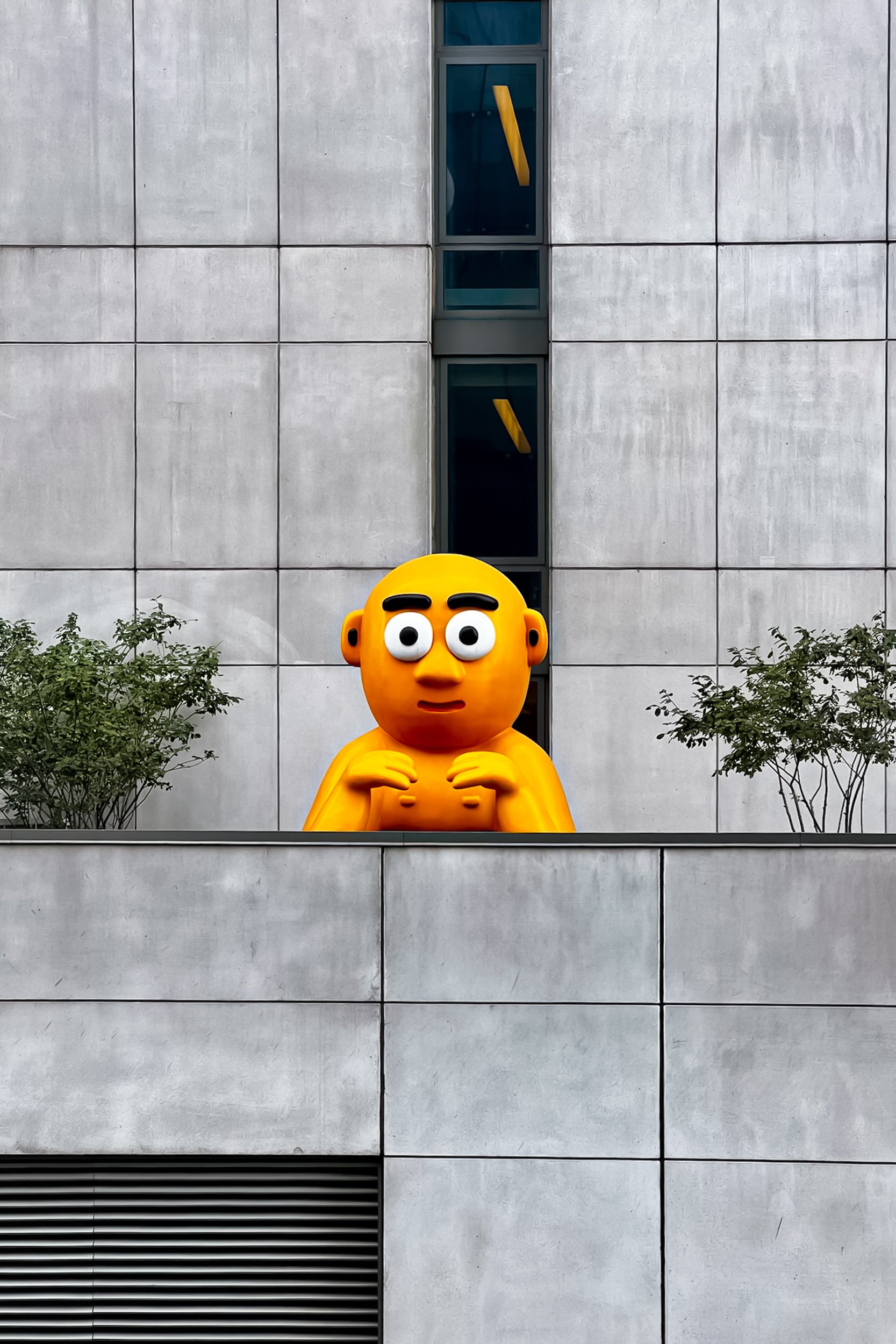 The Lookout - A playful, bright orange-yellow cartoon character sculpture peeking over a concrete balcony ledge, perfectly framed by minimalist grid facade panels and small green bushes.