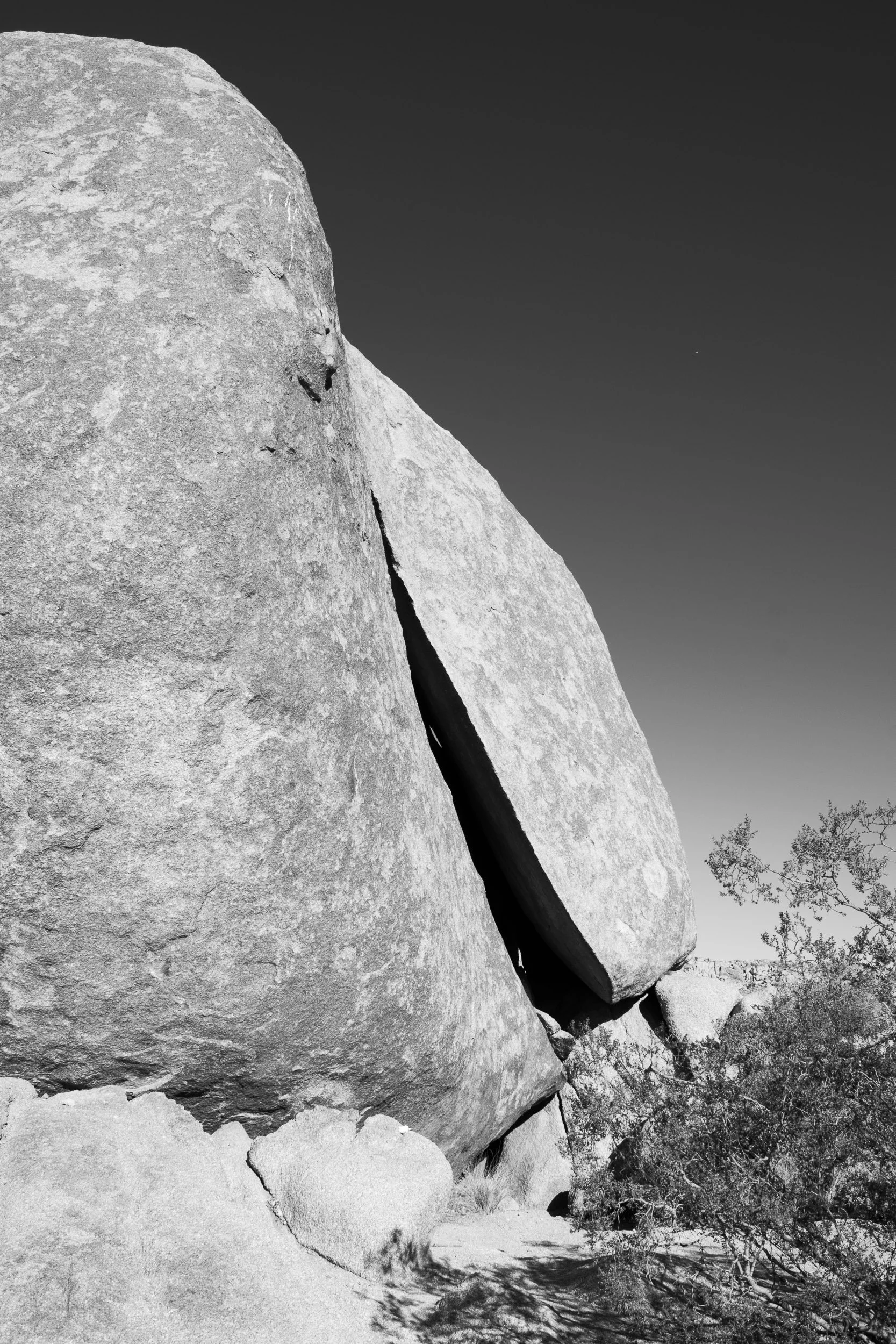 Split Monolith - A towering, high-contrast black-and-white vertical shot of a massive desert boulder cleanly split down the middle, framed by clear skies and sparse desert brush.