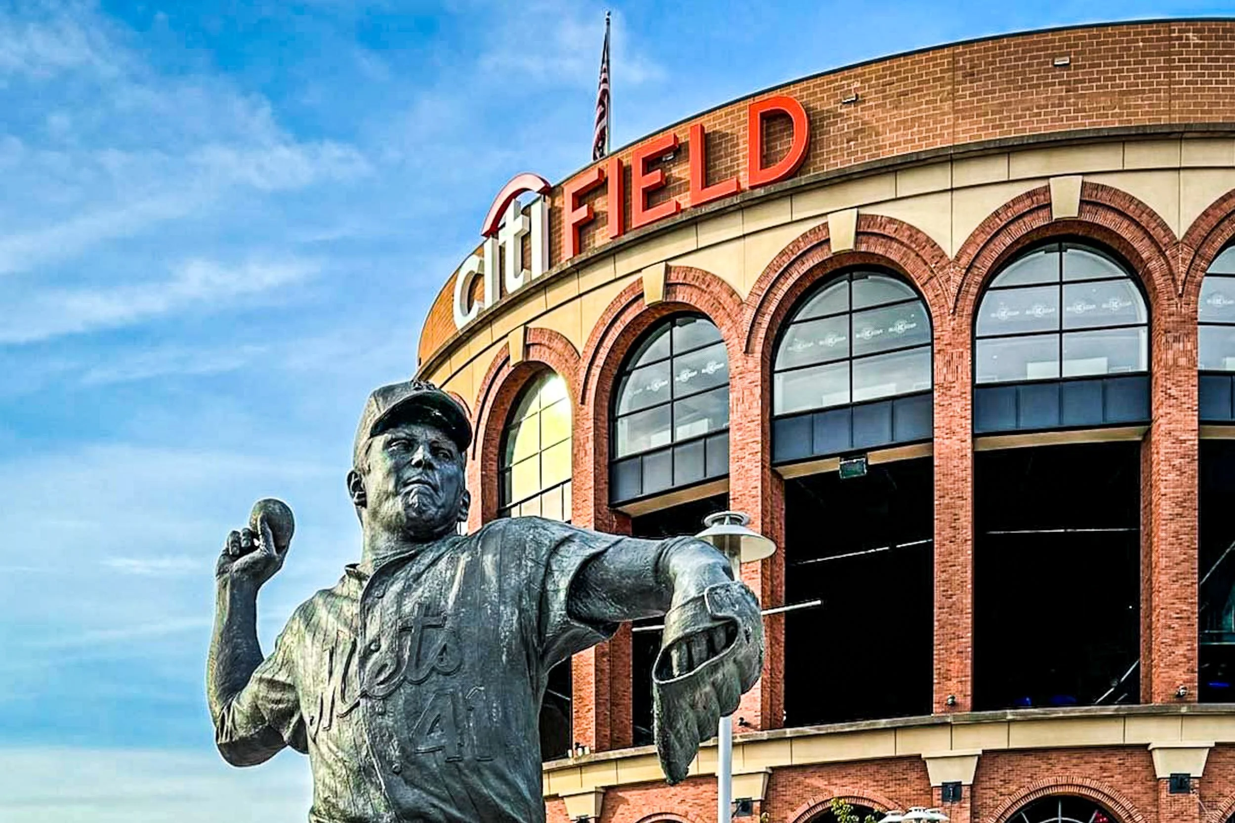 The Franchise - A dramatic, low-angle shot showcasing the iconic bronze statue of Tom Seaver in mid-pitch, proudly framed against the brick arches and bold red lettering of the Citi Field facade under a bright blue sky.