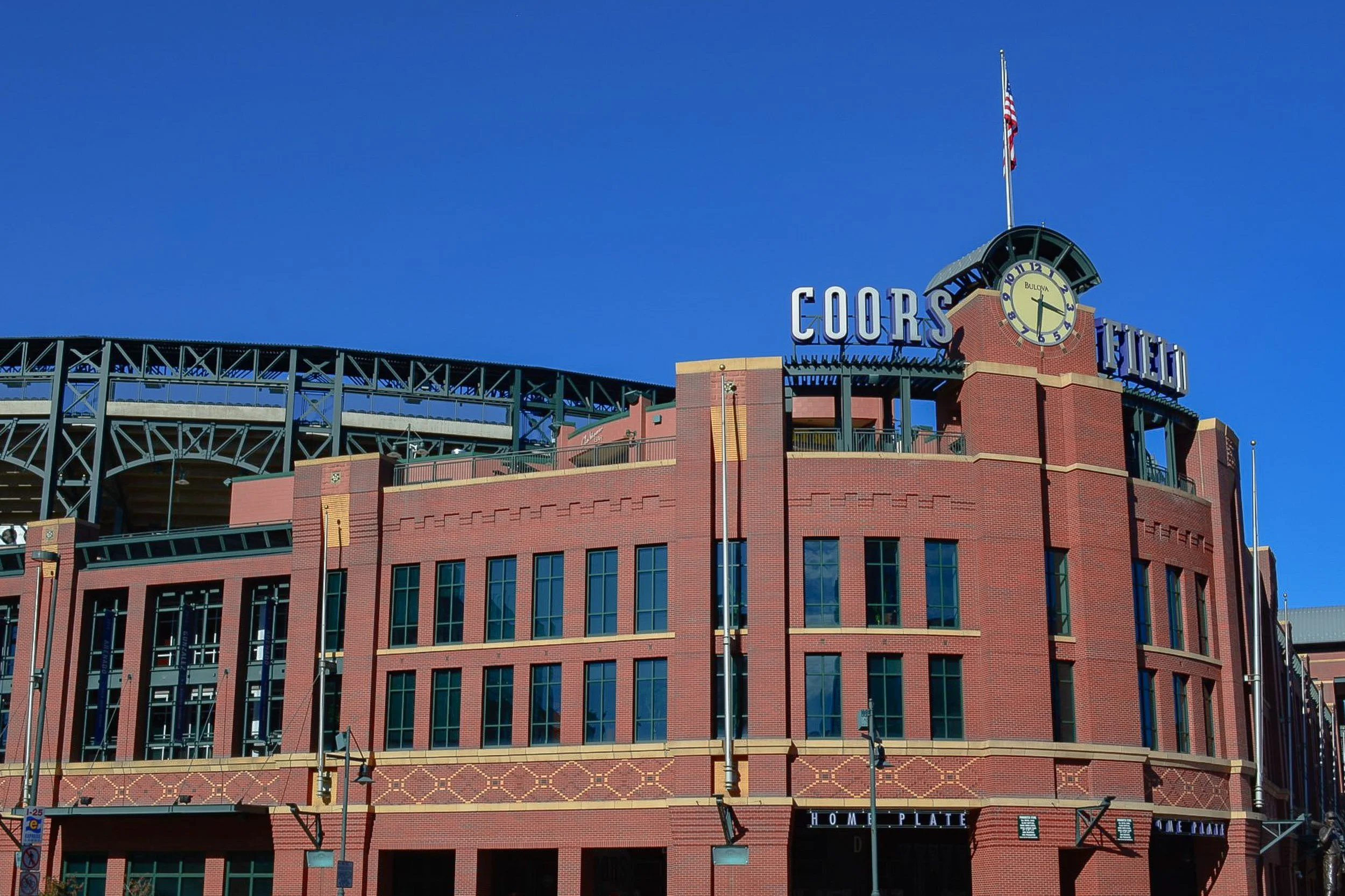 Mile High Brickwork - A wide, architectural landscape capturing the expansive, multi-story red brick exterior and steel framework of Coors Field, highlighted by its prominent corner clock tower against a deep blue sky.