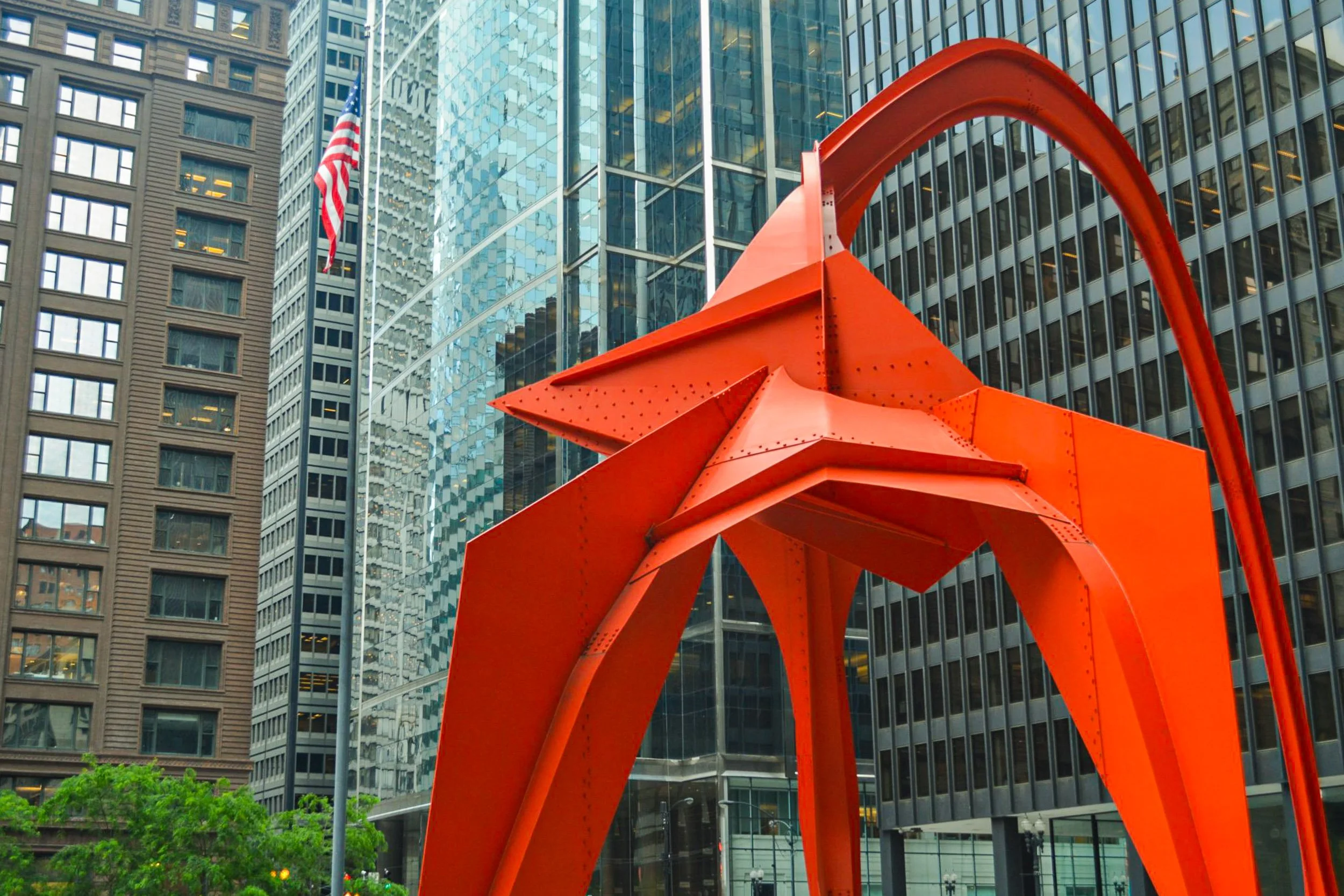 Flamingo Geometry - A vibrant, wide-angle city view focusing on Alexander Calder’s massive, bright red Flamingo stabile sculpture, framed dramatically by the towering glass and steel grids of modern Chicago skyscrapers and a fluttering American flag.