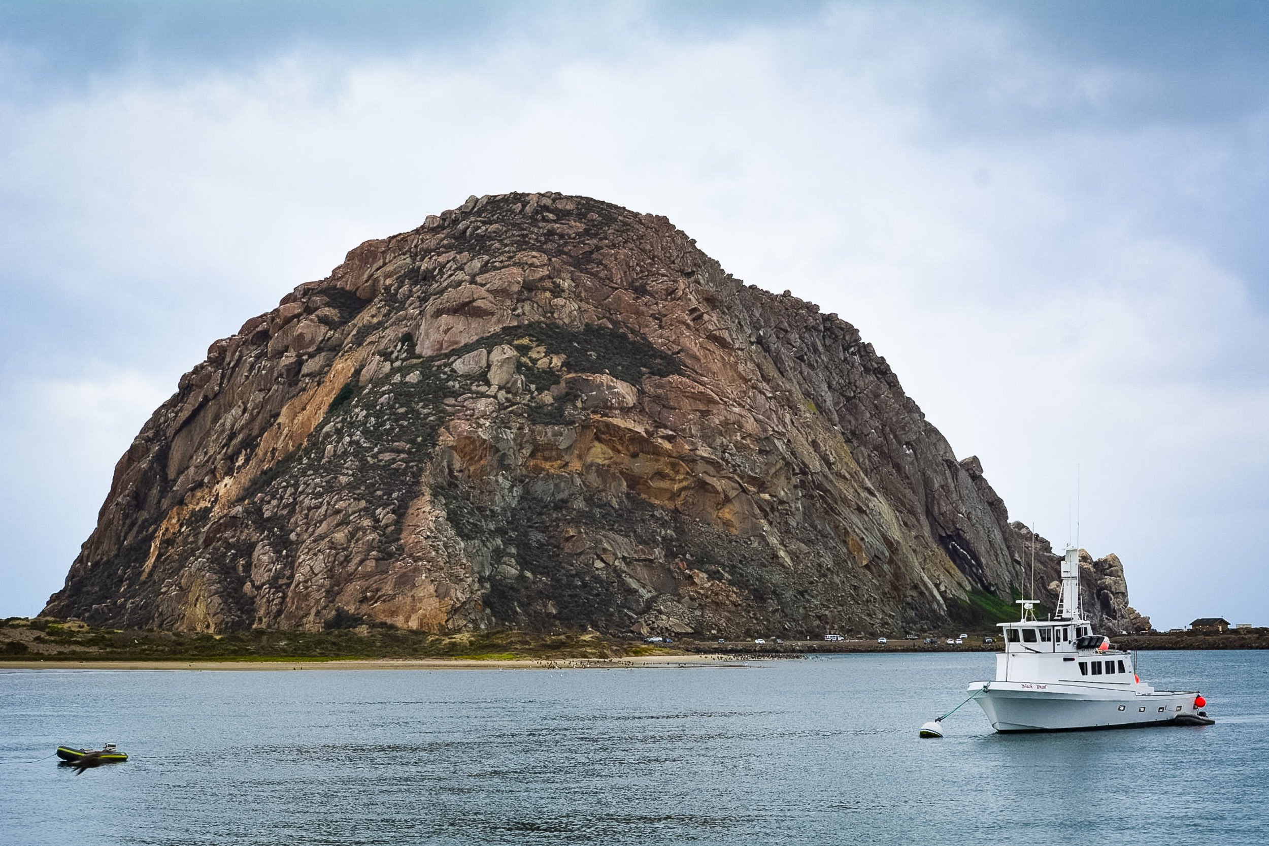 Volcanic Anchor - A wide, scenic coastal landscape featuring the towering, rugged expanse of Morro Rock rising majestically out of calm bay waters, balanced by a clean white motor yacht anchored in the foreground under an overcast sky.