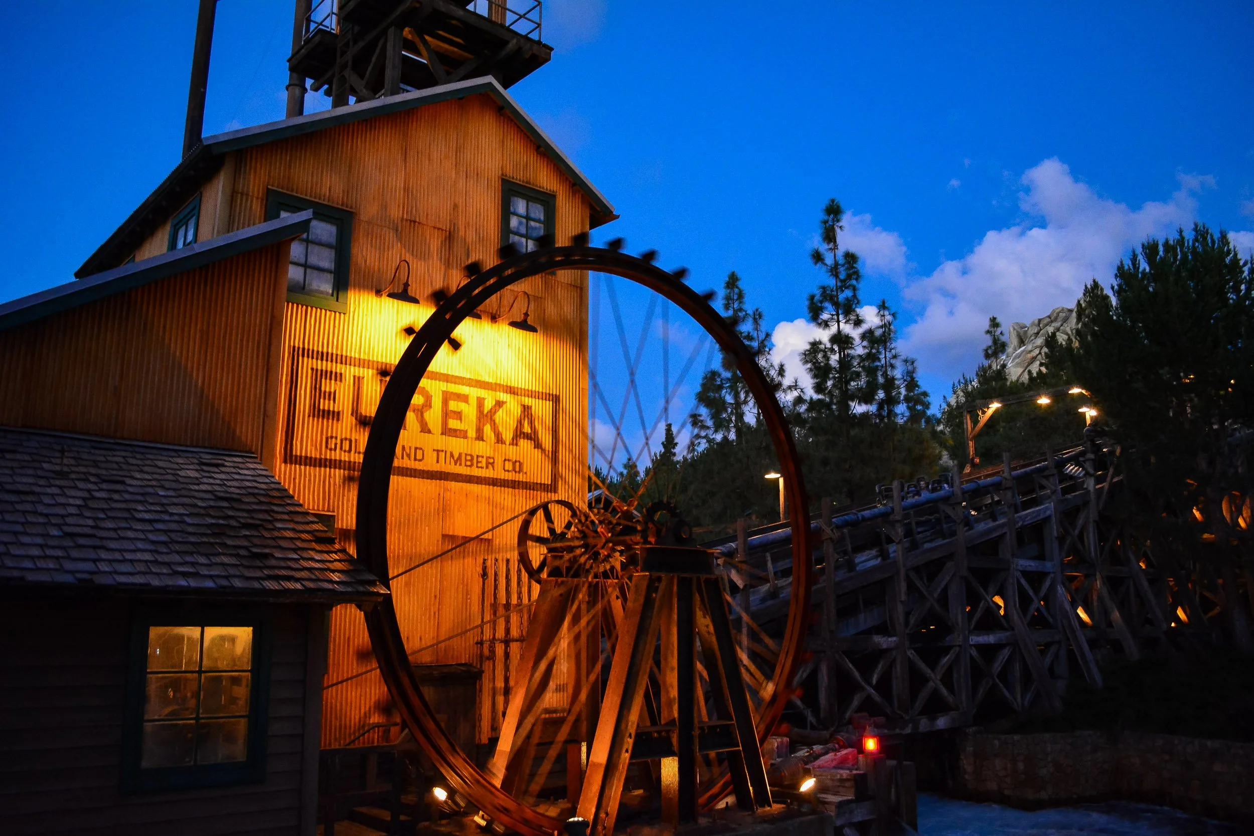 Eureka Mining Motion - A dramatic dusk landscape capturing a large, rustic wooden water wheel spinning with deliberate motion blur outside the illuminated "Eureka Gold and Timber Co." mining building, set against a deep blue evening sky.