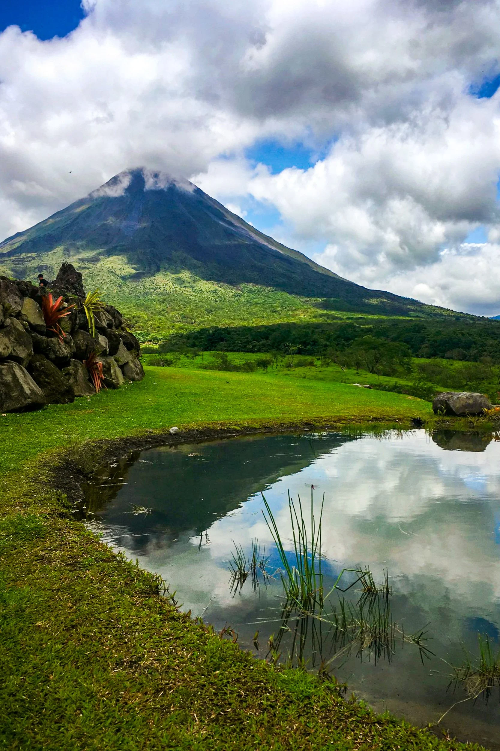 Volcanic Reflection - A prominent volcanic peak shrouded in low clouds, perfectly mirrored in a still pond surrounded by lush green hills.