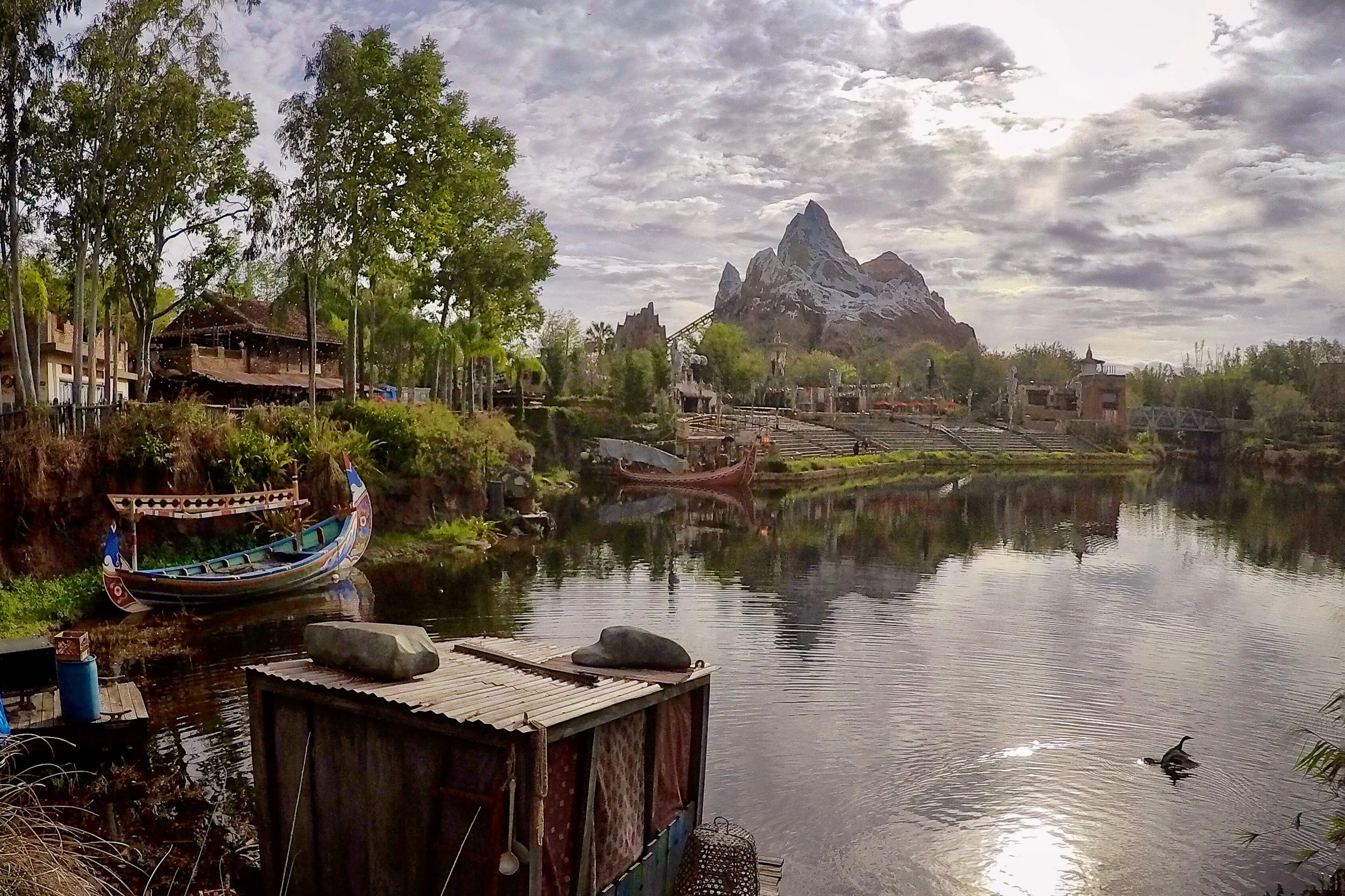 Expedition Waters - A wide, atmospheric landscape capturing a detailed traditional wooden boat moored along a riverbank, with the massive, jagged peak of Expedition Everest reflecting beautifully in the calm water under a dramatic, cloud-filled sky.