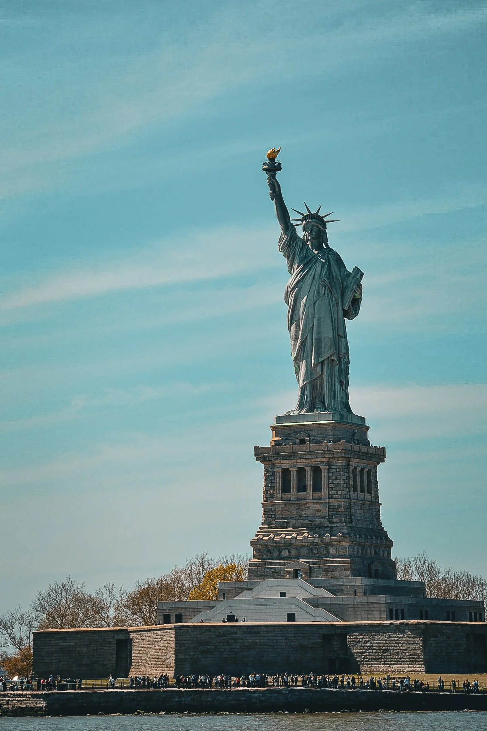 Lady Liberty - A classic, full-length profile shot of the Statue of Liberty rising majestically against a soft, bright blue sky with wispy clouds.