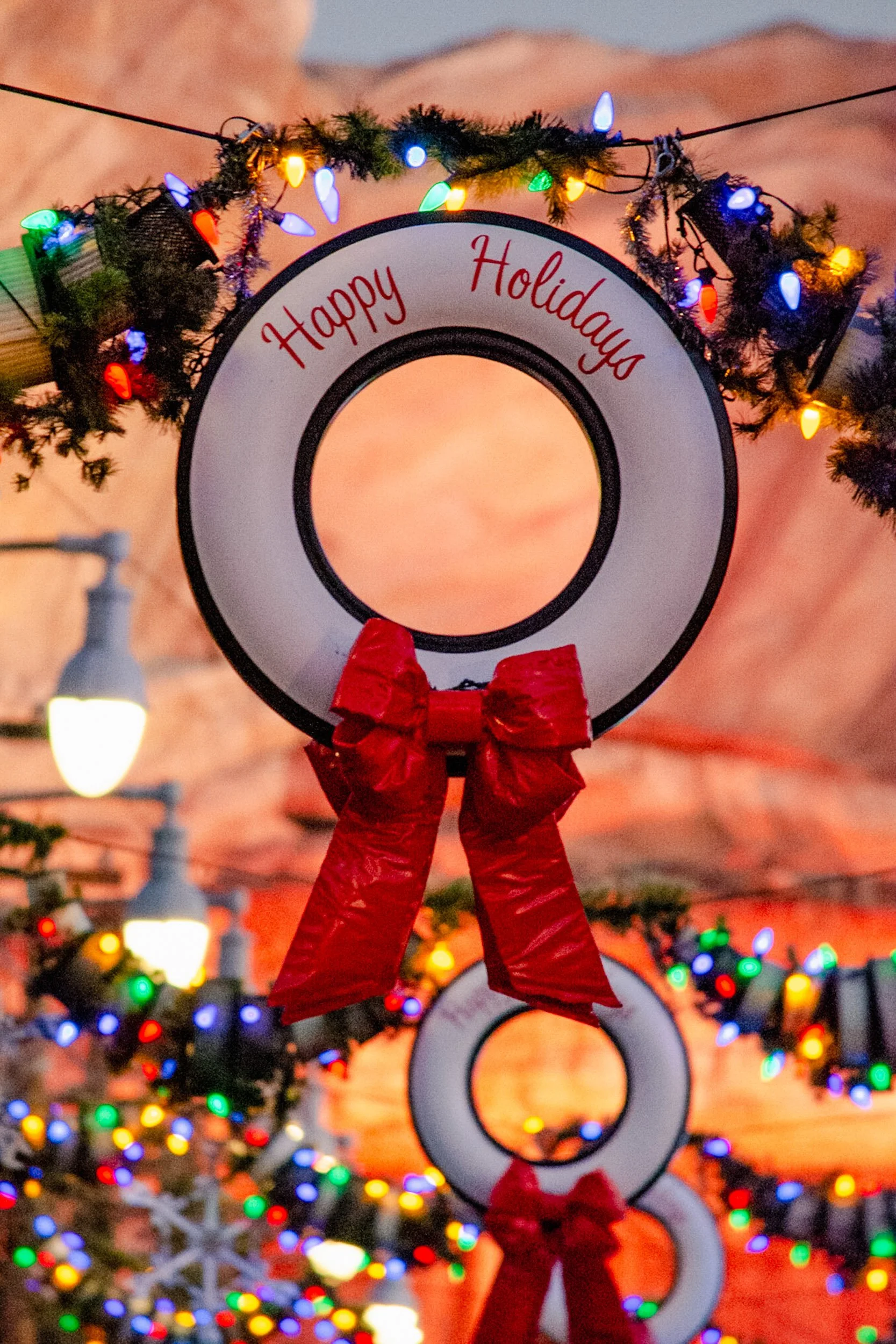 Radiator Springs Cheer - A festive close-up focusing on a unique holiday wreath crafted from a classic white-walled car tire and a bright red bow, draped in colorful Christmas string lights with warm desert cliffs blurred in the background.