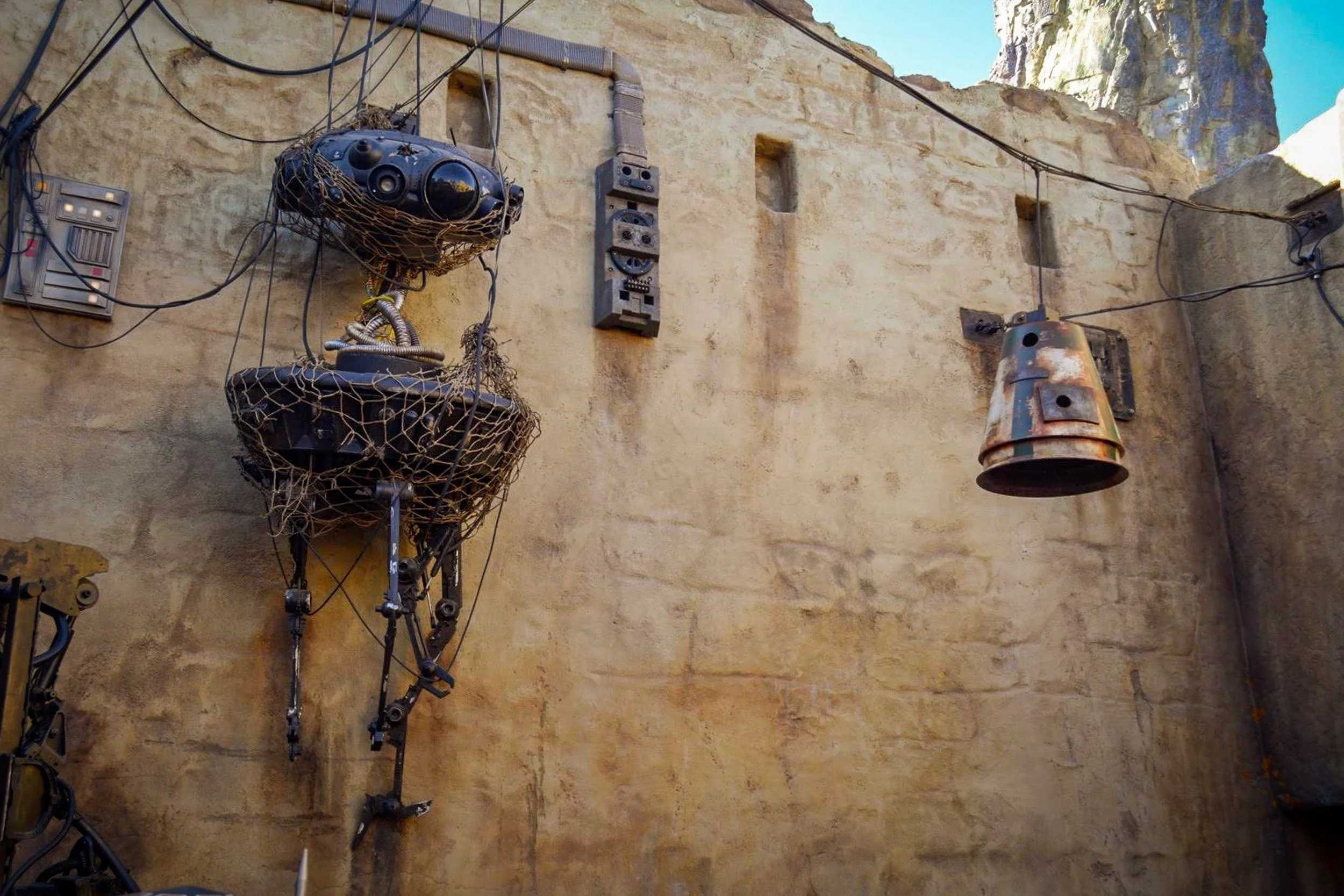 Outpost Junk - A textured, industrial details shot featuring a deactivated, skeletal black droid hanging from messy overhead wiring against a weathered, sun-baked mud wall in Galaxy's Edge.
