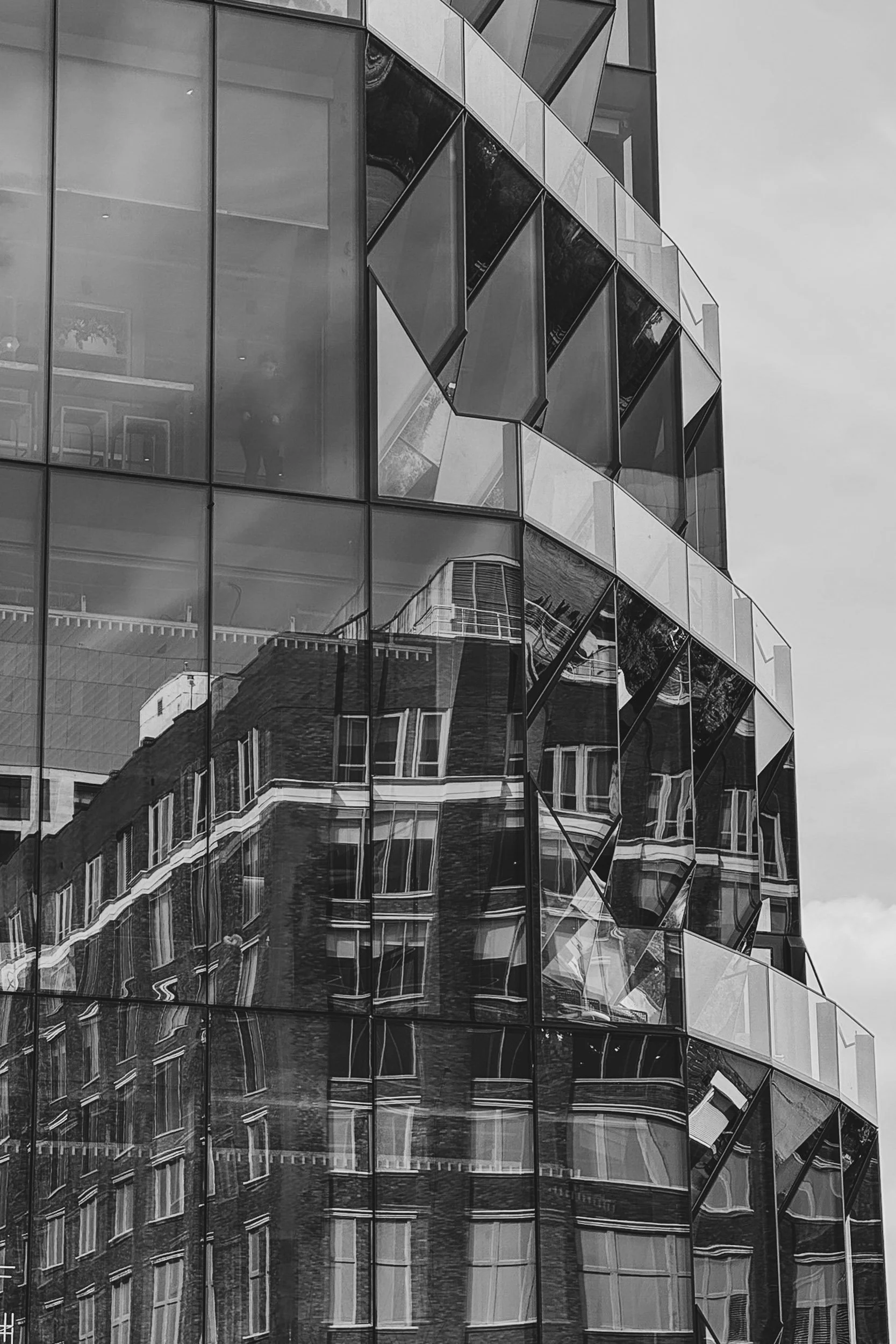 Fractured Facade - A striking, high-contrast black-and-white vertical photograph of a modern glass high-rise, capturing complex, geometric distortions of an older brick building reflected across its multi-angled windows.