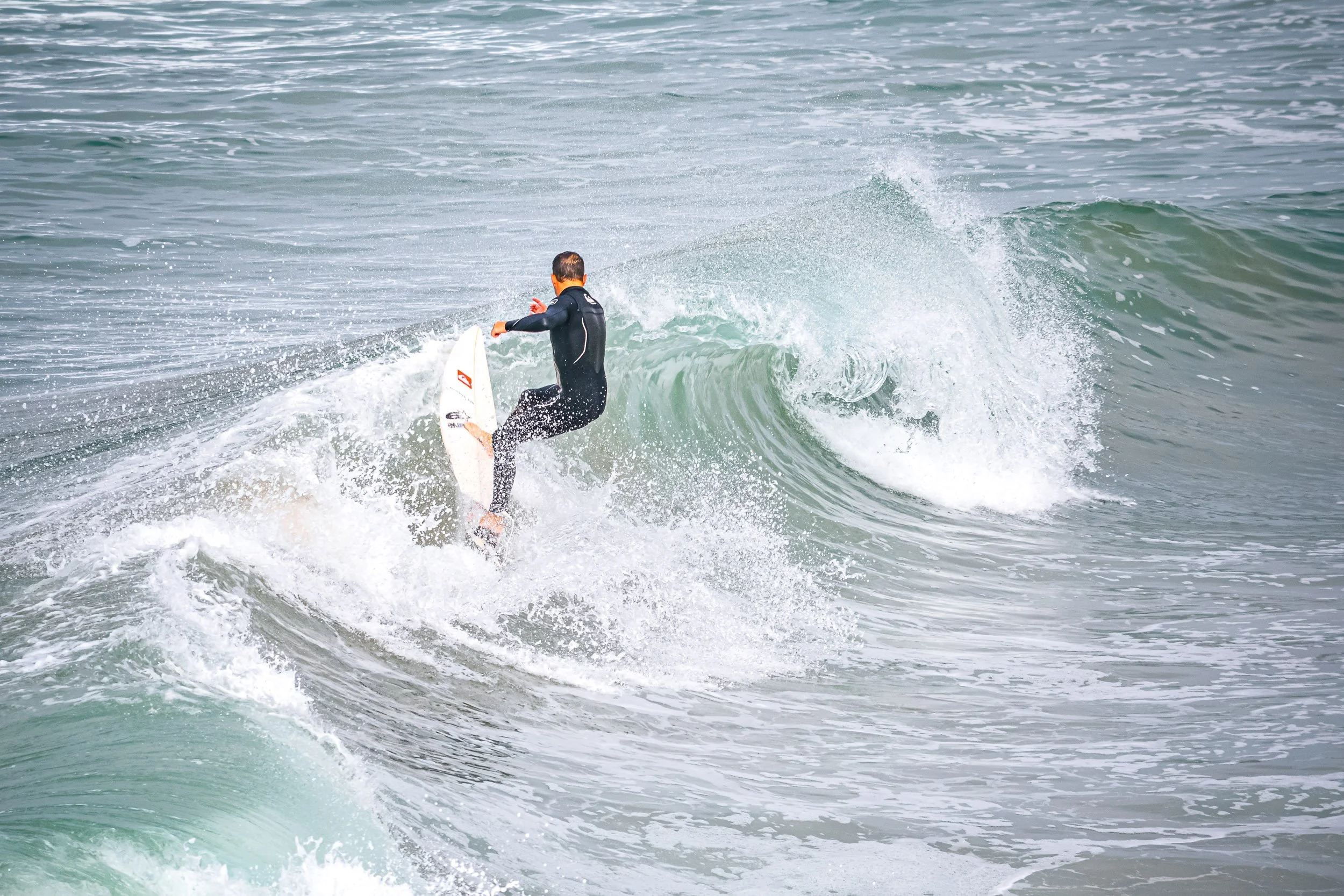 Crest of the Wave - An action-packed sports photograph tracking a surfer in a black wetsuit skillfully carving across the glassy green face of a breaking ocean wave, surrounded by spraying white foam and churning surf.
